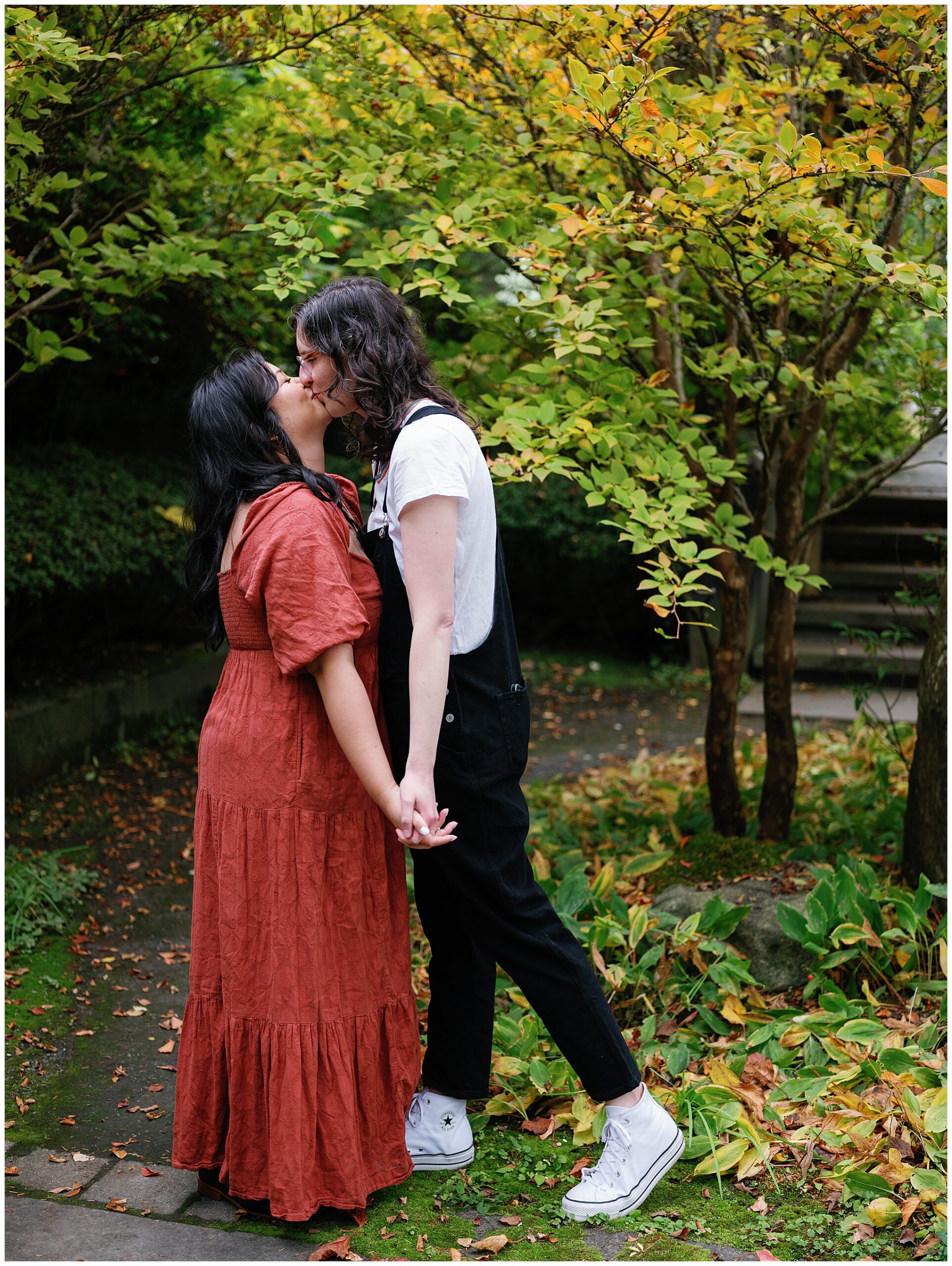 Carli and Erin kissing while holding hands in a garden.