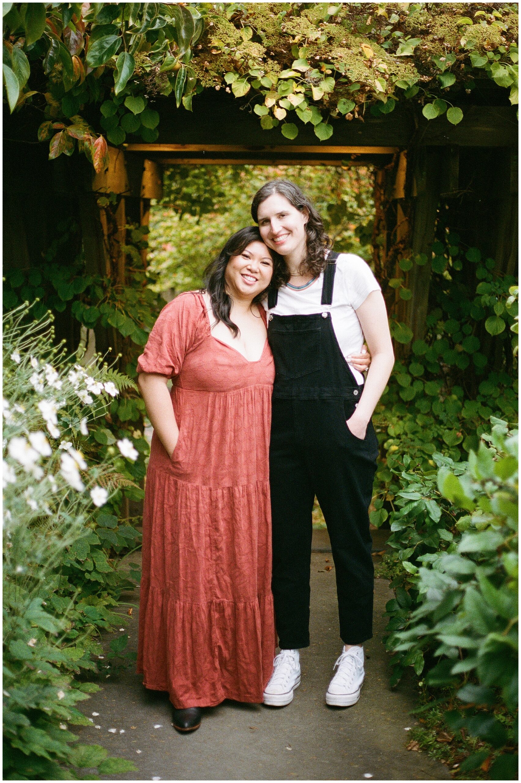 Carli and Erin stand arm-in-arm under a garden archway, smiling sweetly together.