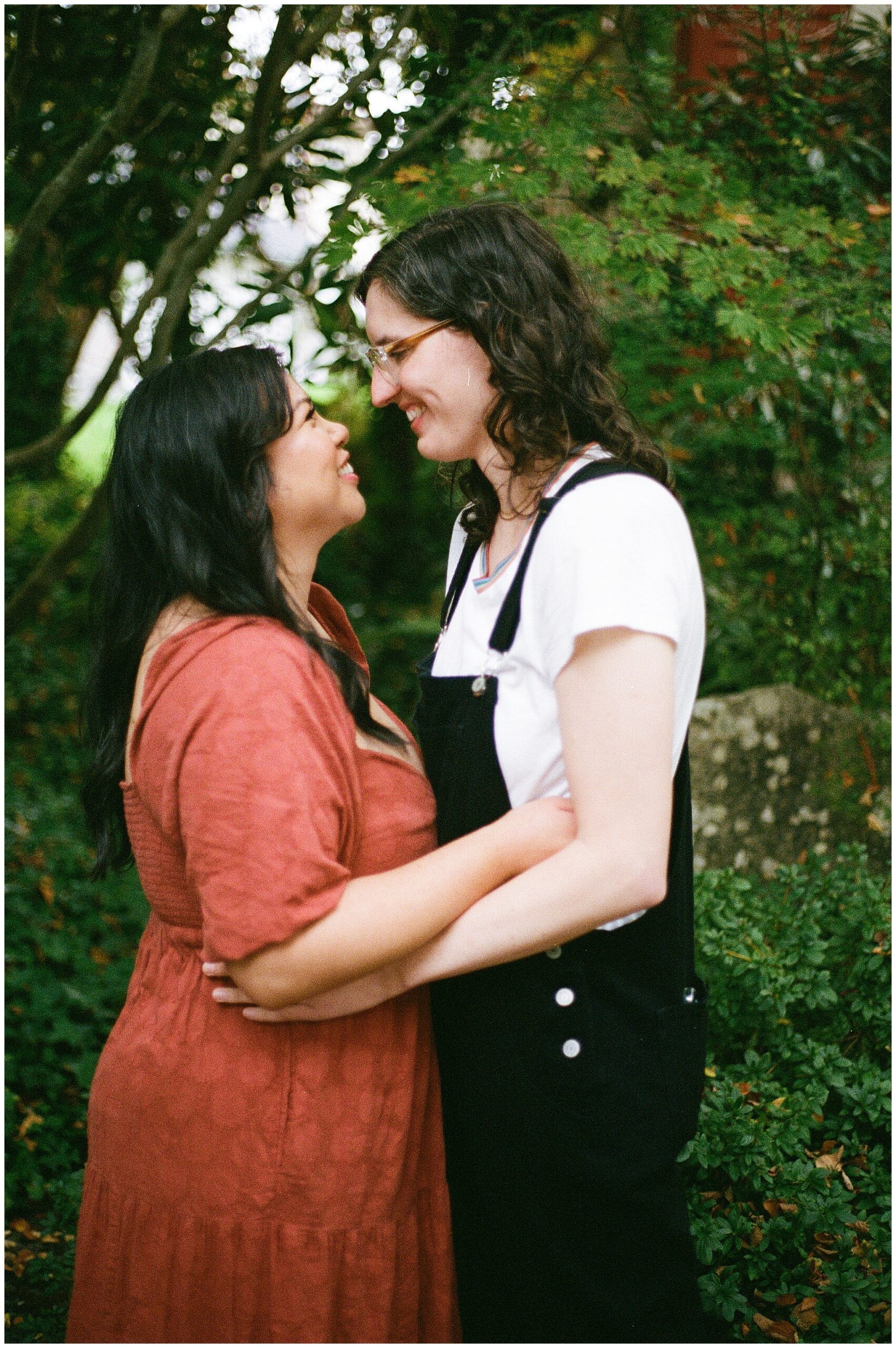 Carli and Erin embrace, smiling warmly at each other in a lush garden.