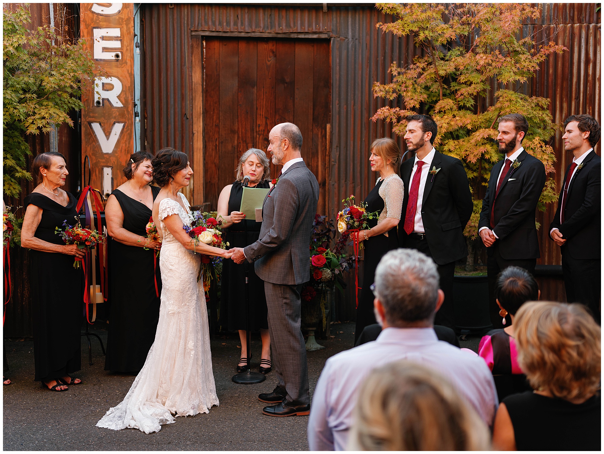 Couple exchanging vows surrounded by their wedding party under a rustic sign.