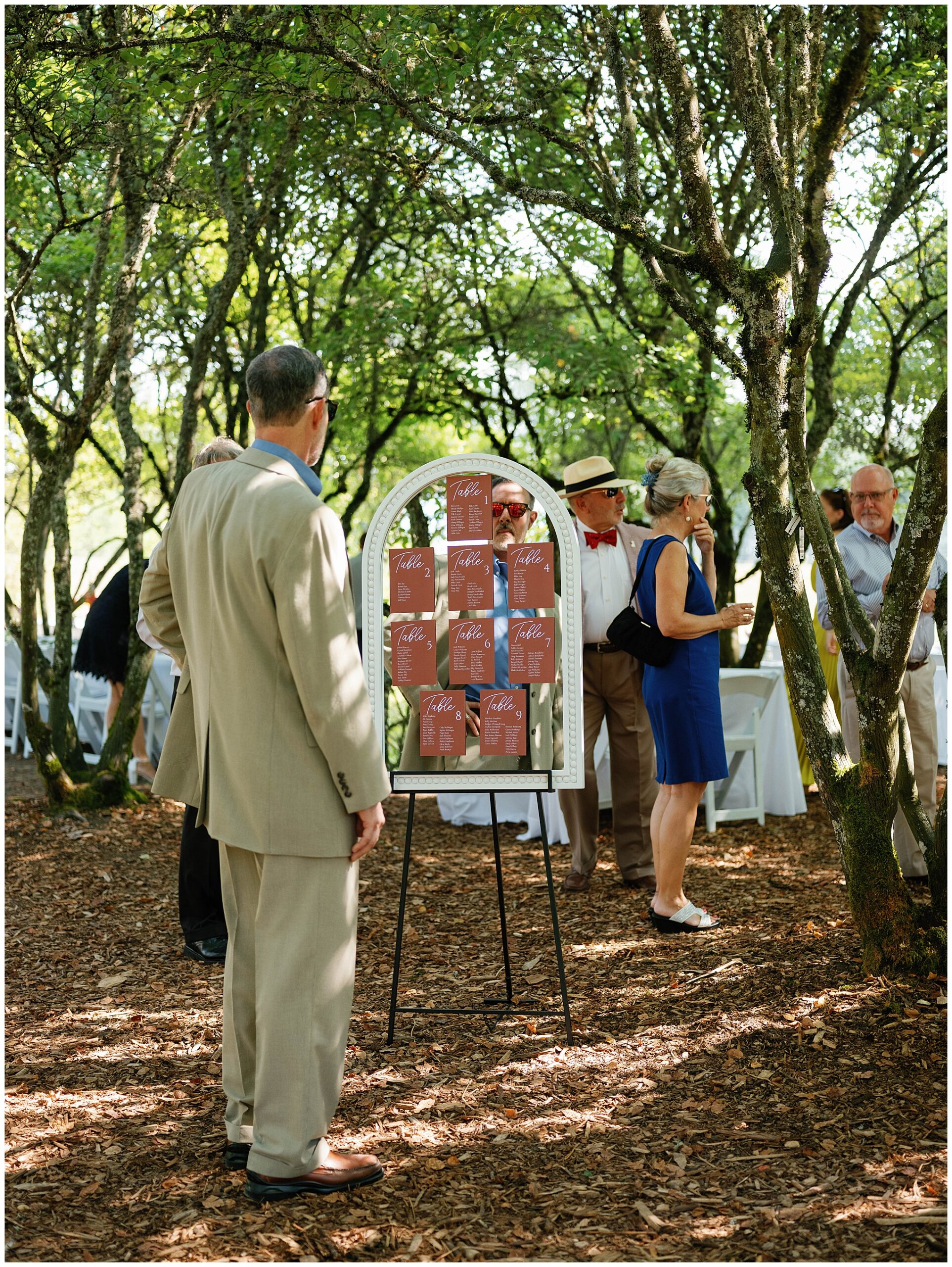 Guests gather around the seating chart displayed in a forested area during Grace and Chris's wedding at the Center for Urban Horticulture.