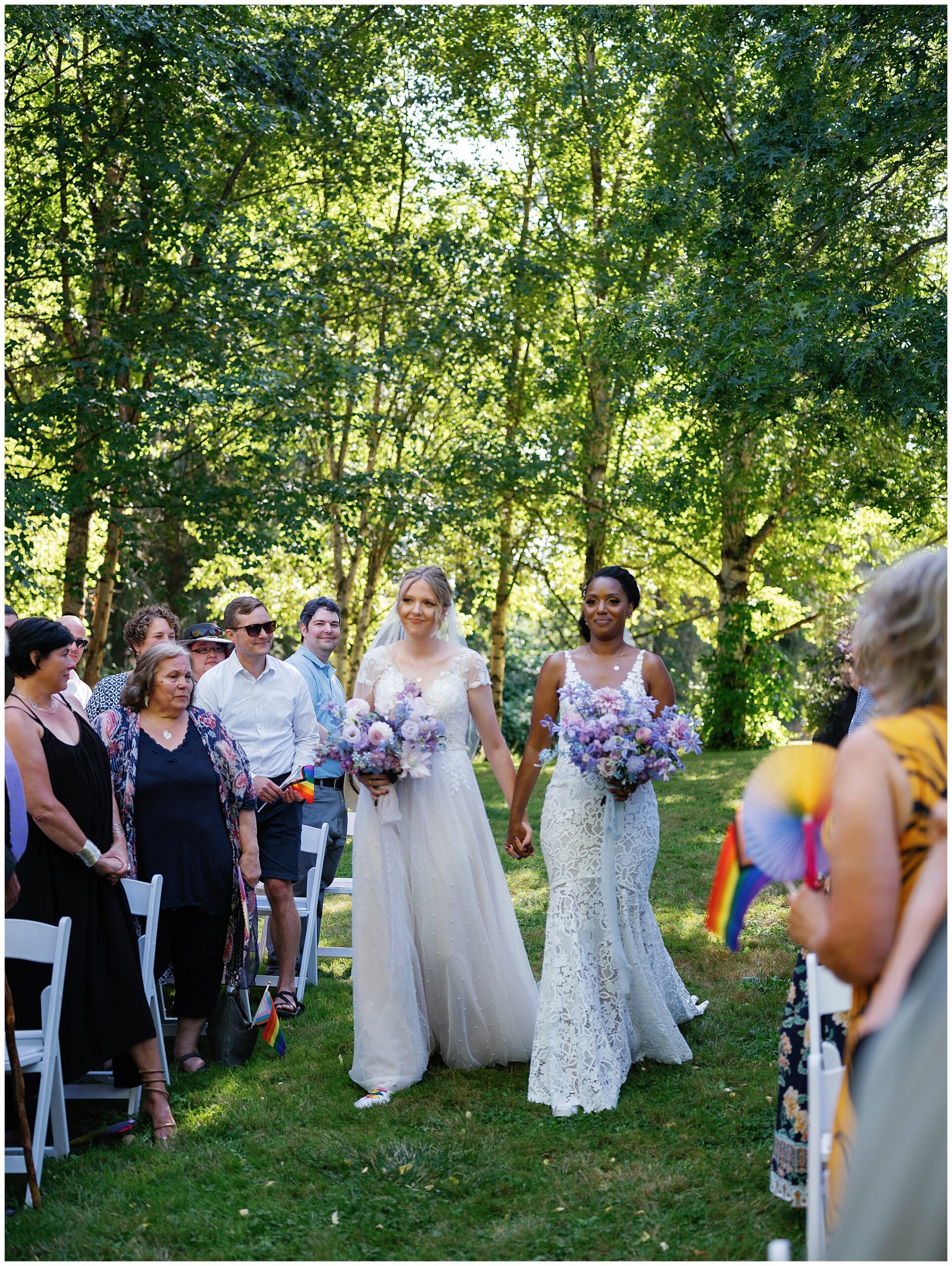 The brides walking down the aisle hand-in-hand, surrounded by guests, amidst a lush garden setting.