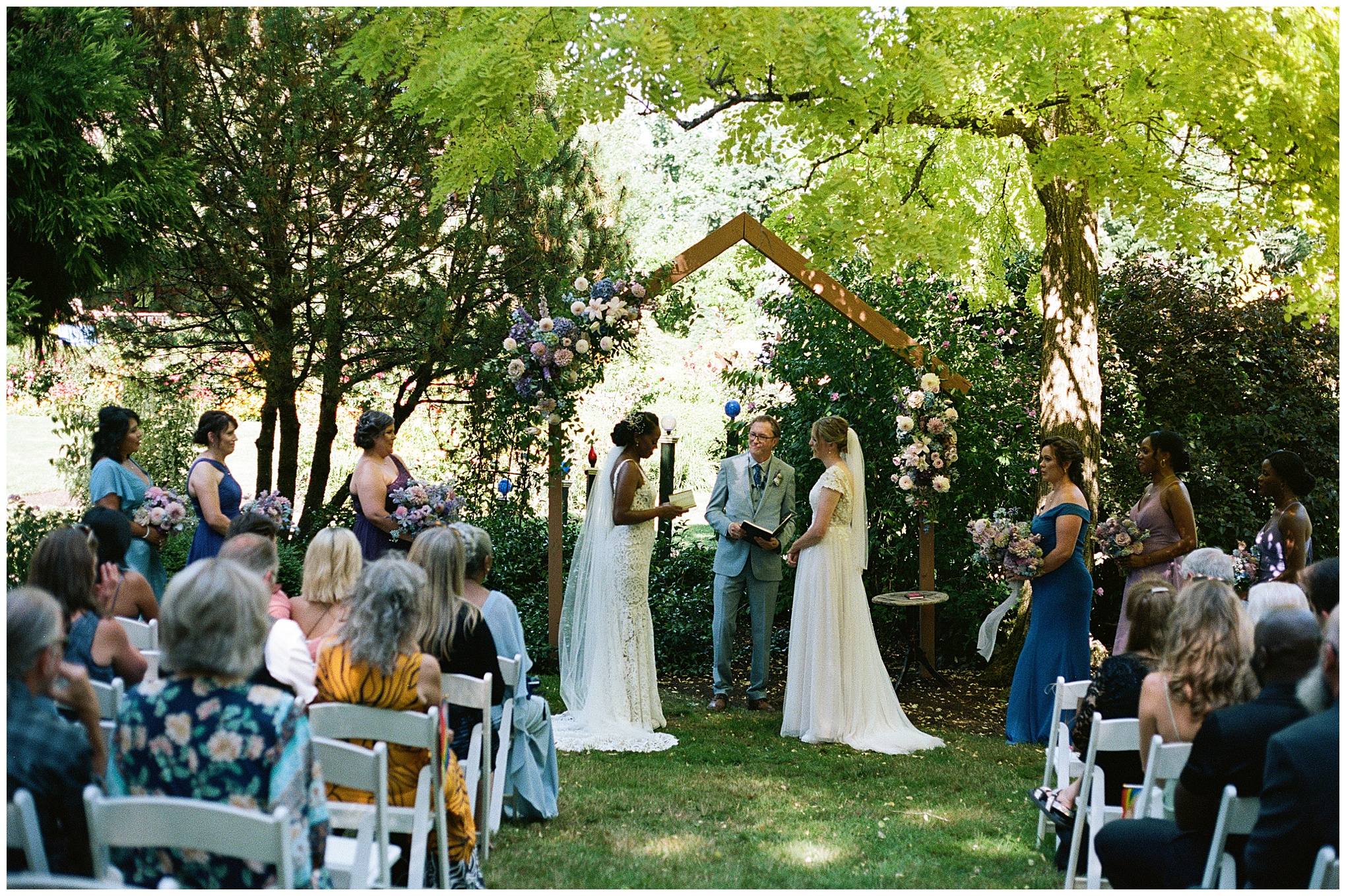 The couple exchanging vows under a wooden arch adorned with flowers, surrounded by bridesmaids and guests.