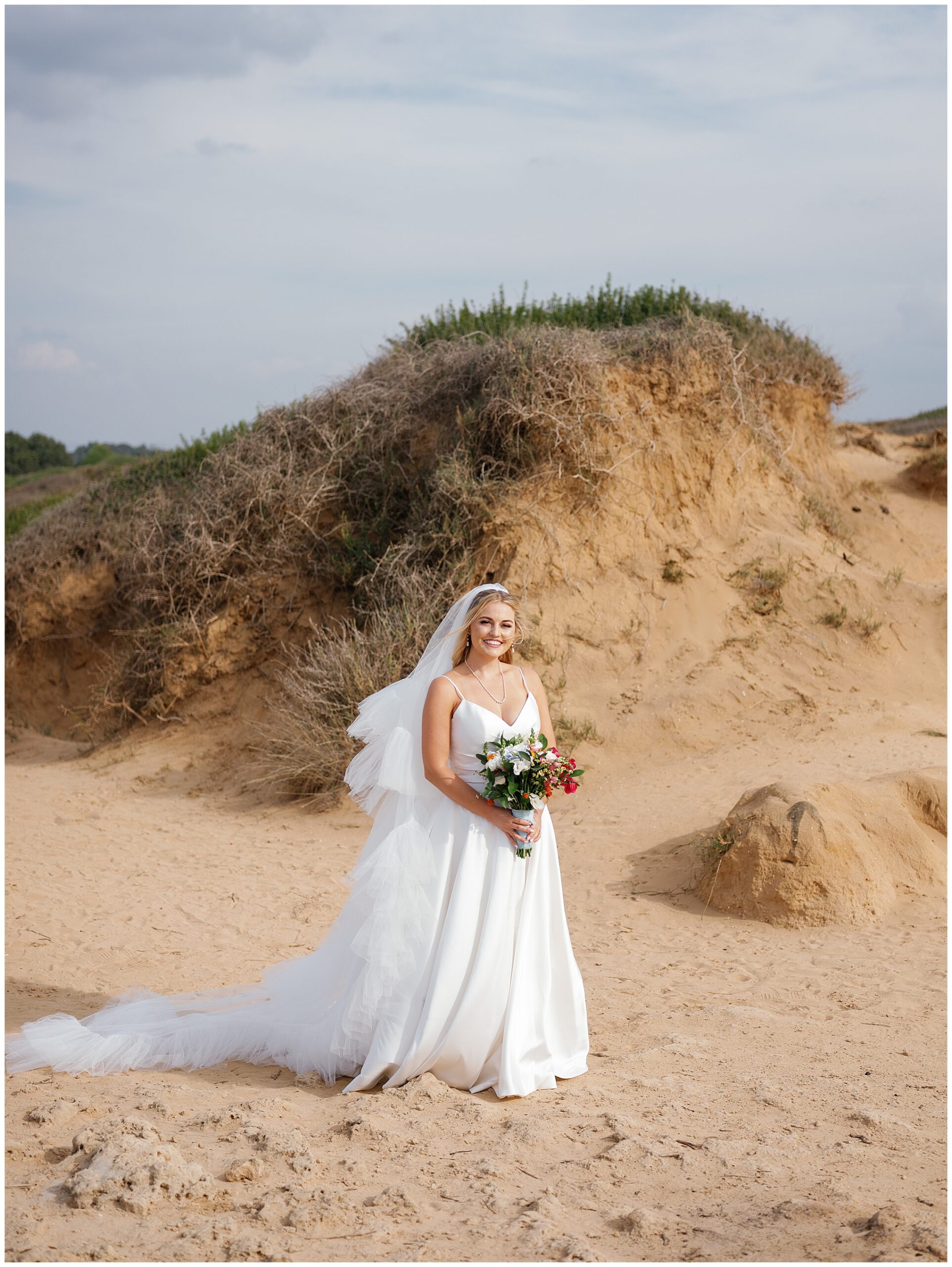 Amelia standing on the sand in her flowing wedding dress, holding a bouquet, with soft cliffs in the background.