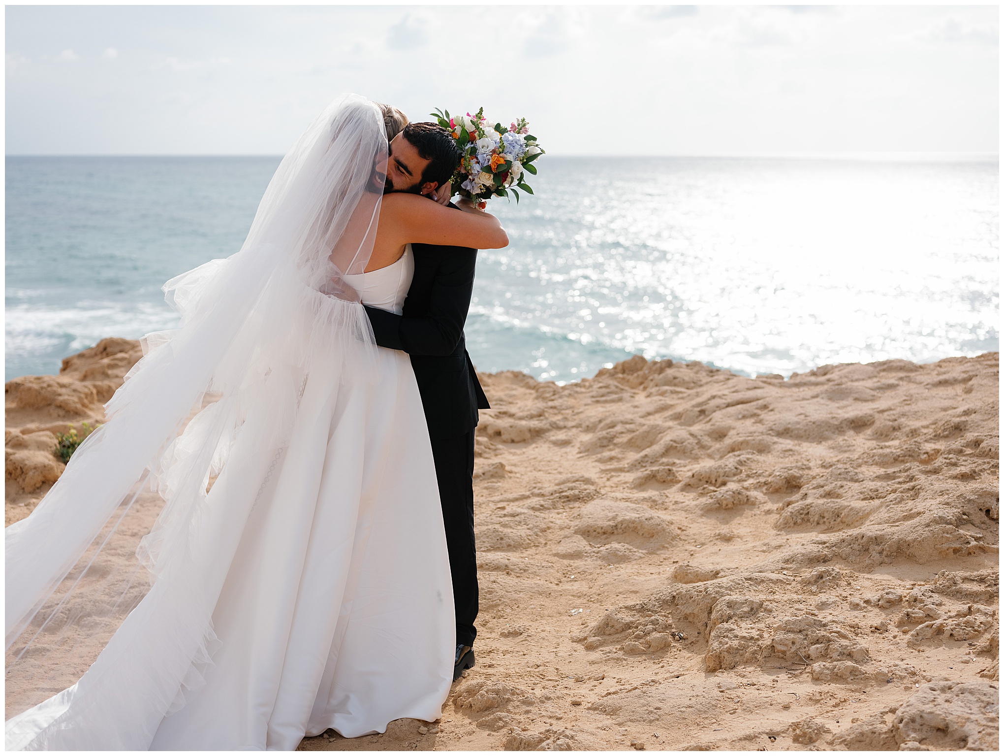 Bride and groom embrace during their cliffside wedding, overlooking the Mediterranean Sea as sunlight reflects on the water.