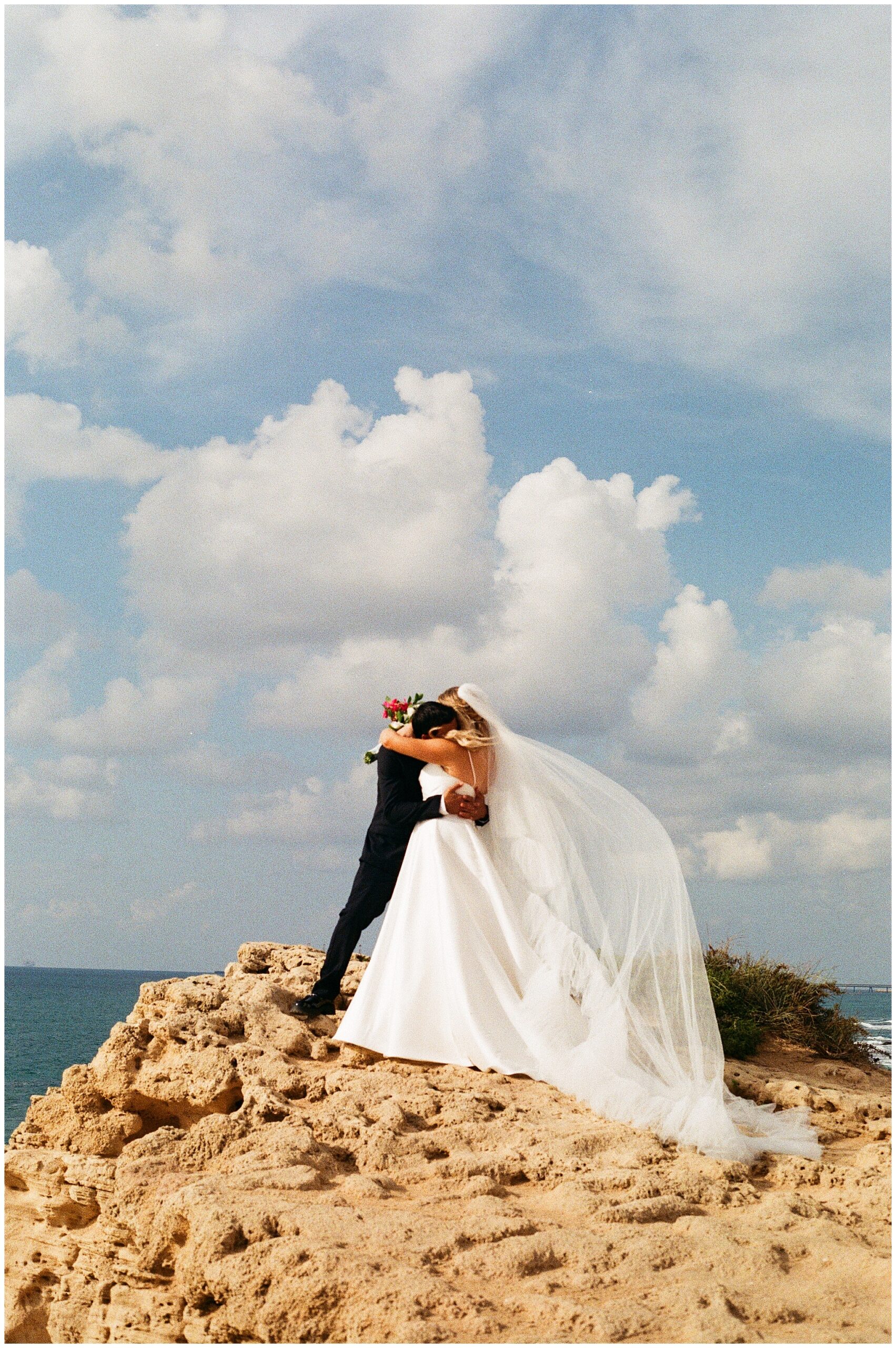 Bride and groom hug on a rocky cliffside with dramatic clouds and the ocean in the distance.