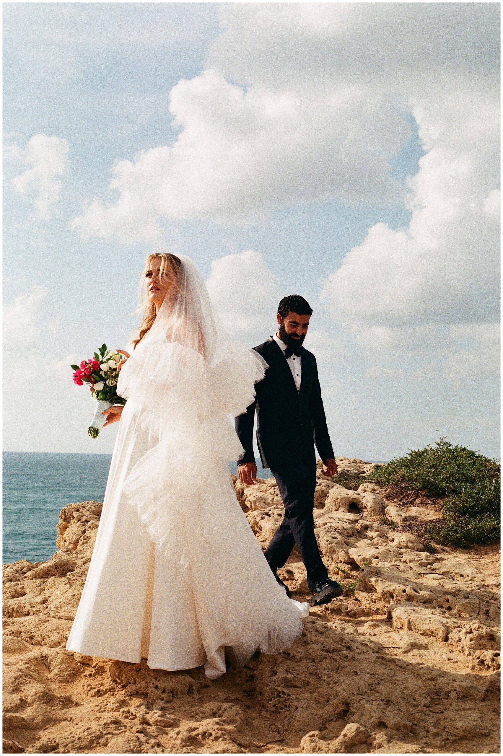 Bride holding a bouquet and groom walking together along the edge of a cliff overlooking the Mediterranean Sea.