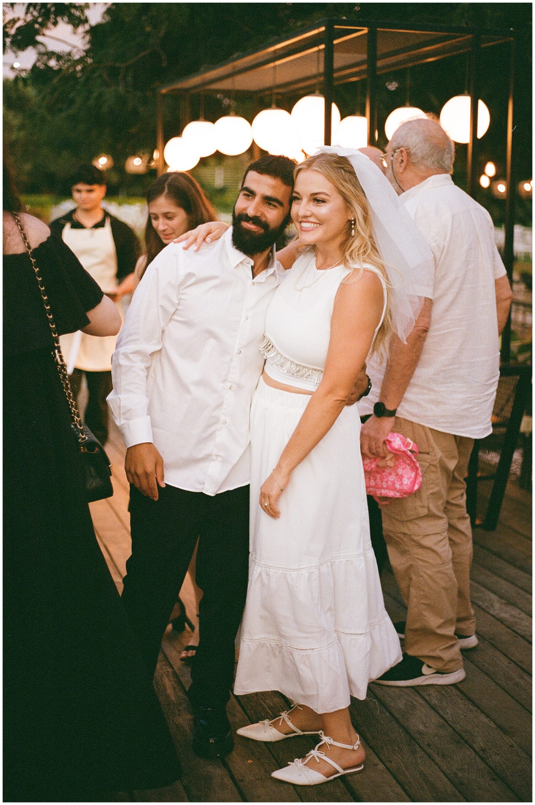 Bride and groom smile together during their evening wedding reception, surrounded by guests and soft lighting.