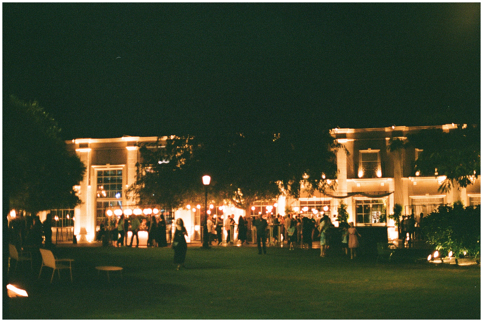 Nighttime view of wedding venue with illuminated building and guests mingling outside during Amelia and Lotan’s wedding.