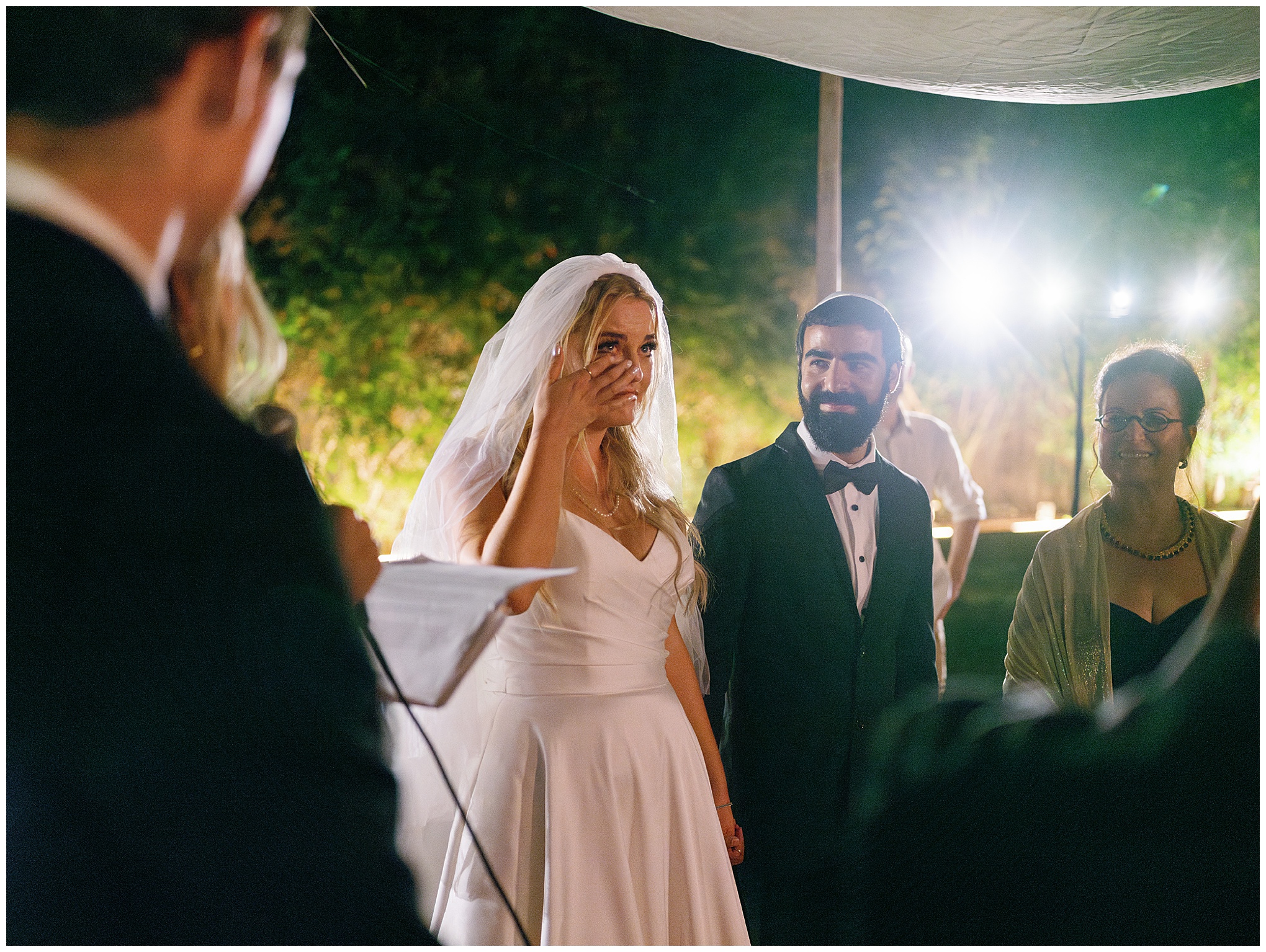 Bride wipes away tears during an emotional moment in the wedding ceremony, standing with the groom under soft lighting.