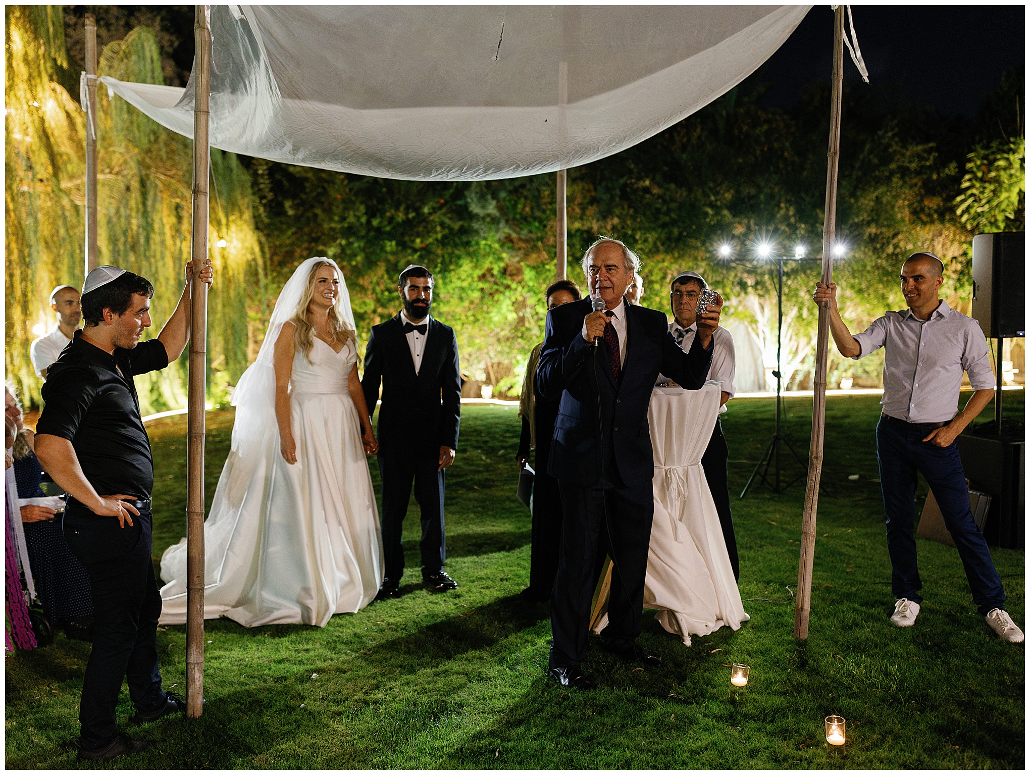 Wedding ceremony under the chuppah with a speaker addressing the bride, groom, and guests in an outdoor evening setting.