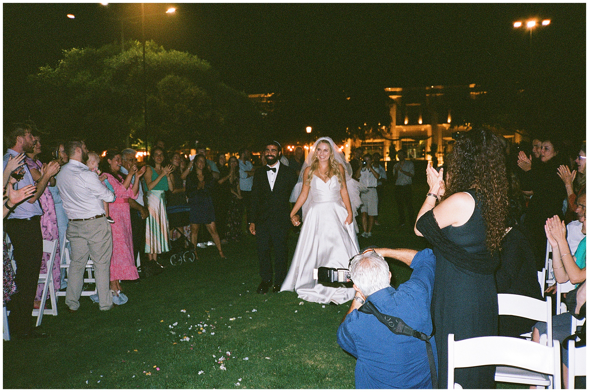 The bride and groom hold hands as they walk down the aisle at night, surrounded by applauding guests.