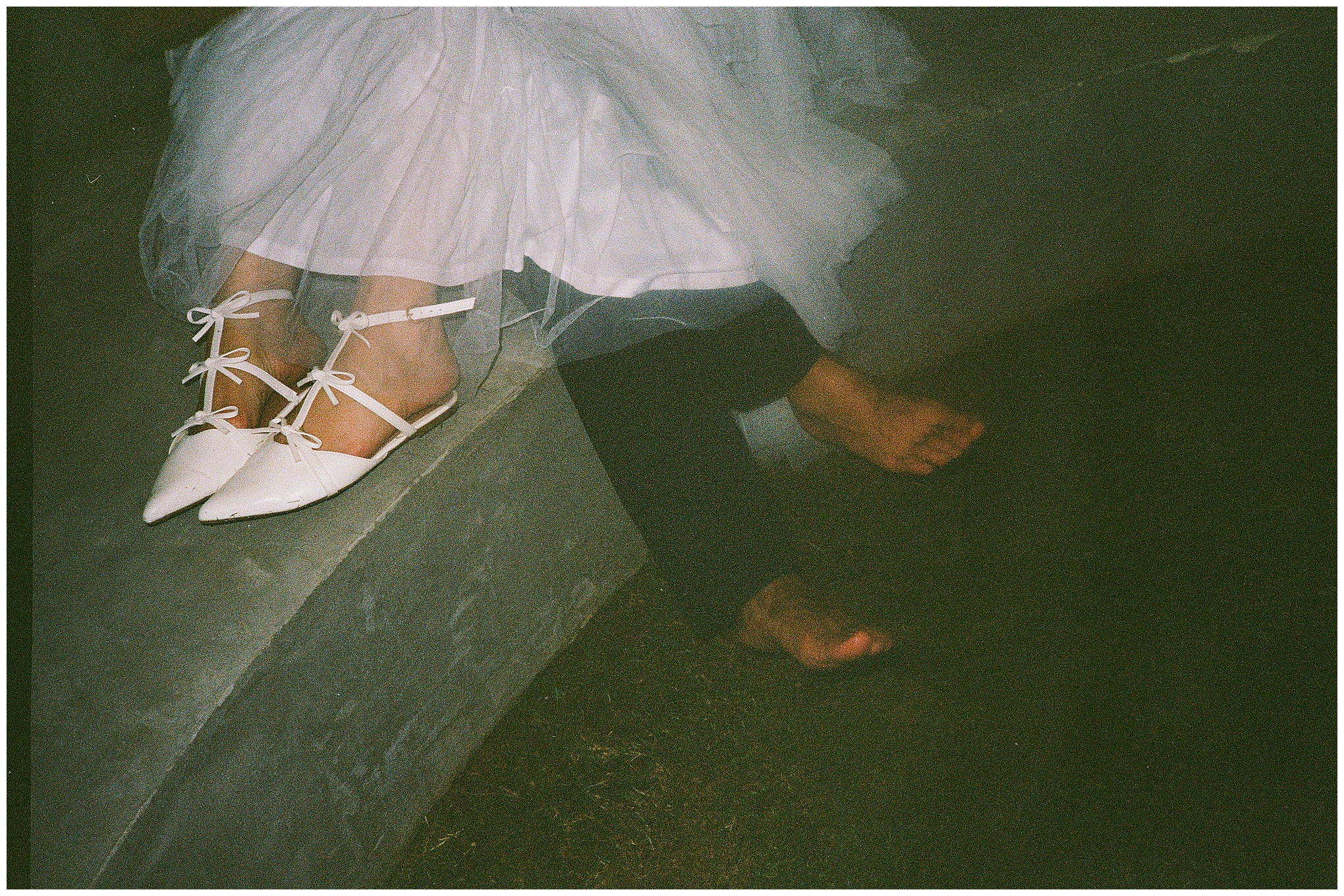 The bride’s white pointed shoes and the groom’s bare feet rest on a concrete ledge.