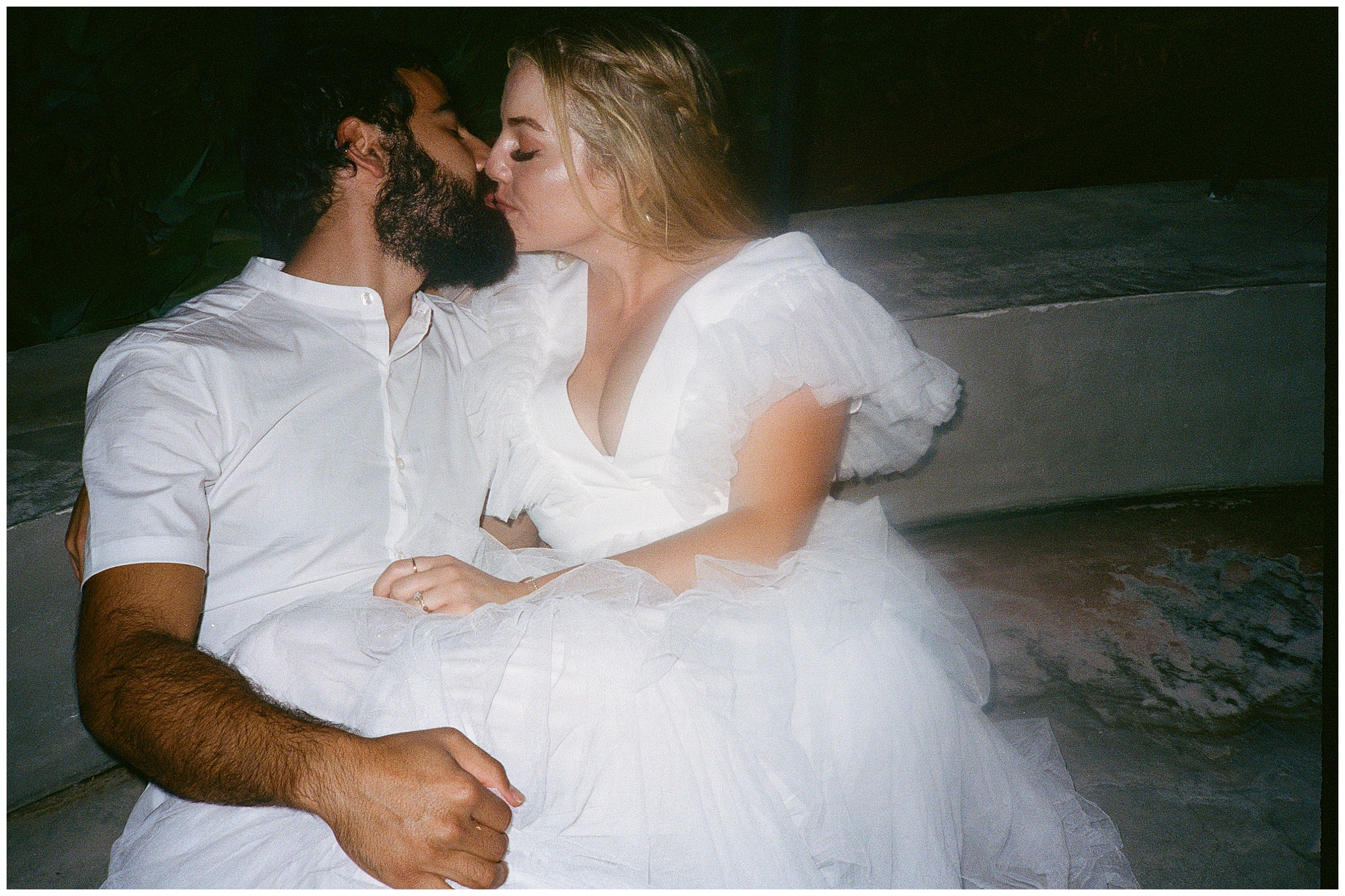 The bride and groom share a kiss while sitting on a ledge, enjoying a quiet moment together after their wedding.