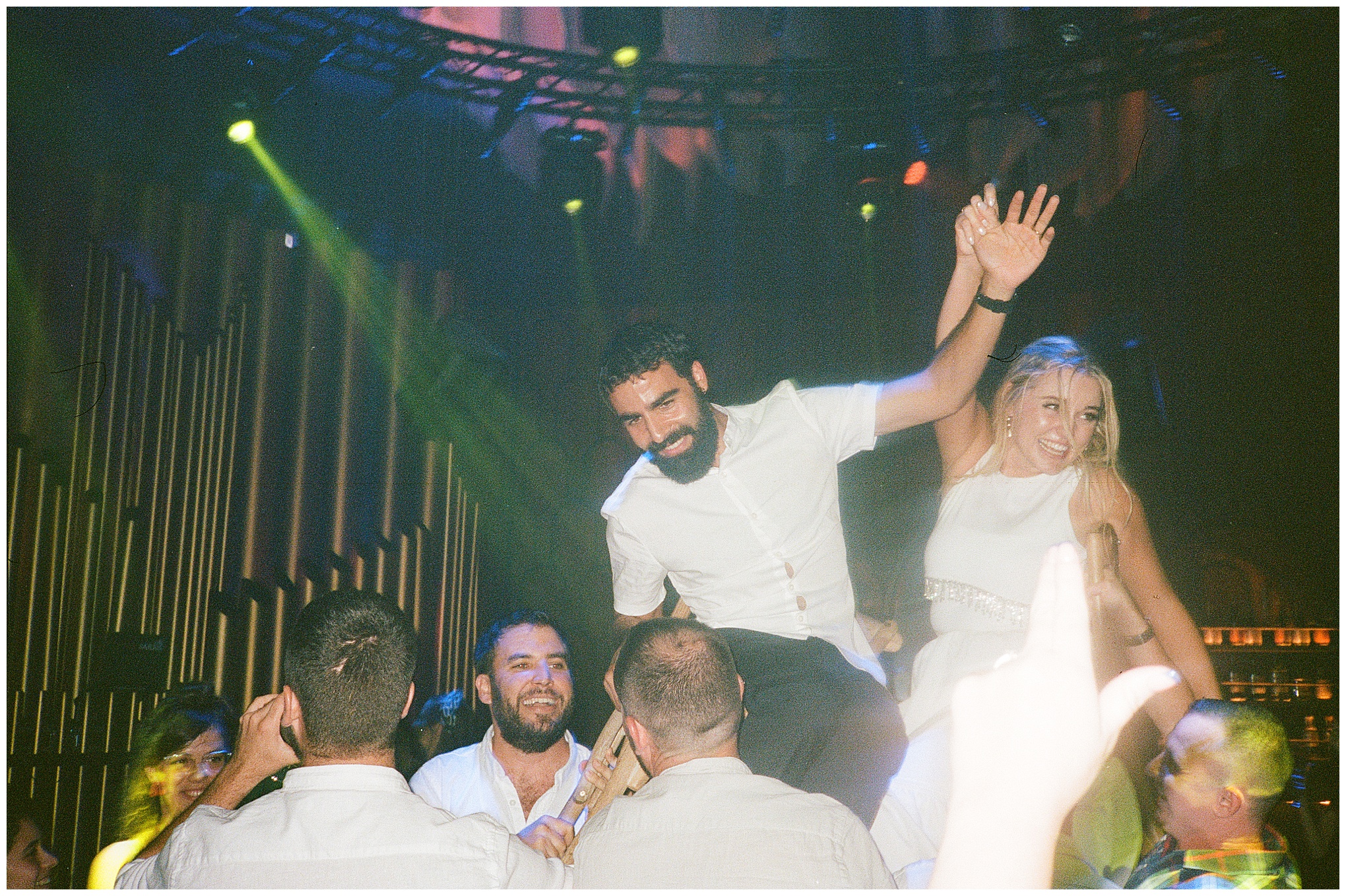 The bride and groom are lifted on chairs during a lively reception dance surrounded by cheering guests.