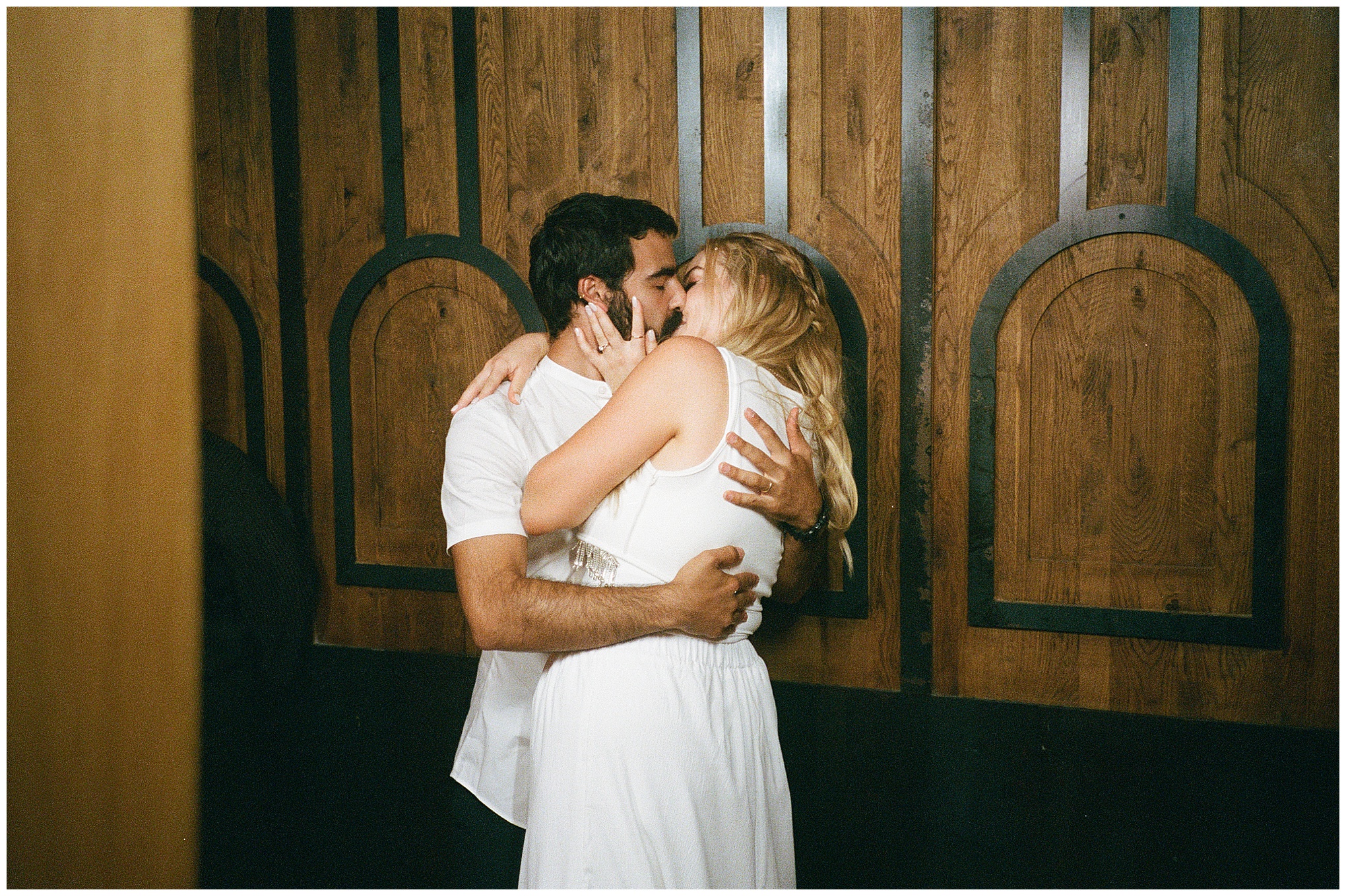 The bride and groom embrace and kiss against a wooden wall during a quiet moment at their wedding reception.