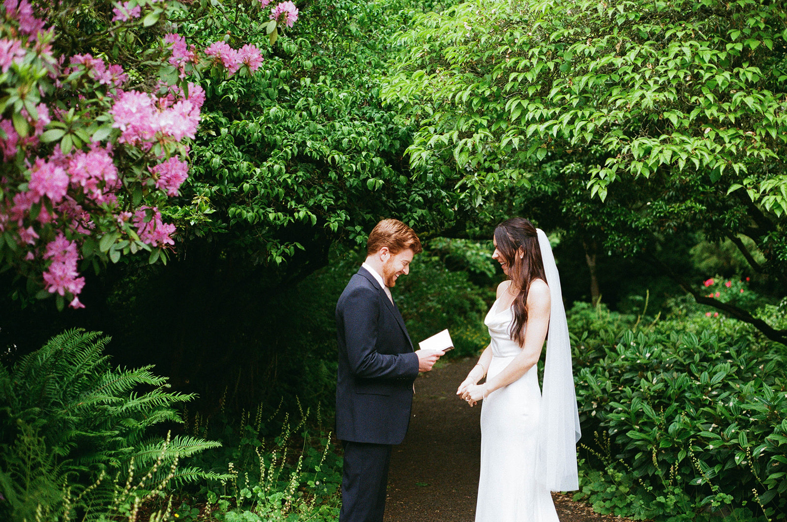 Groom reading his vows to the bride in a lush garden surrounded by greenery and pink blossoms at Parsons Gardens.