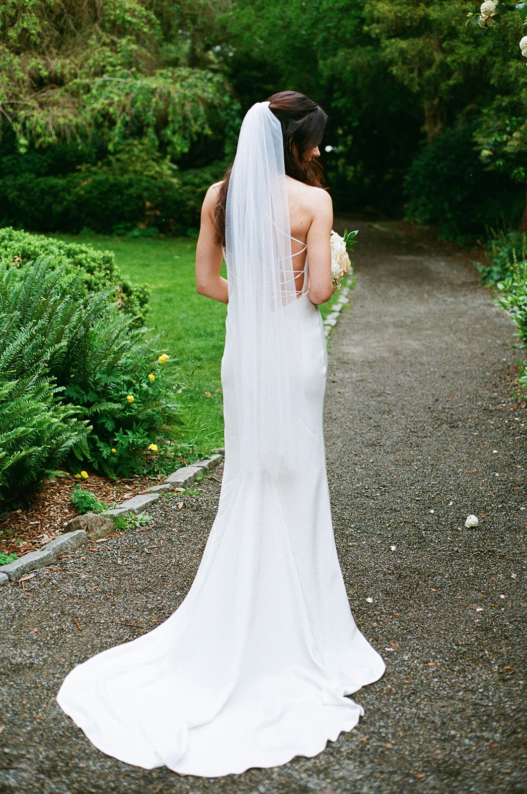 Bride standing in a garden with her back to the camera, wearing a long veil and elegant gown at Parsons Gardens.