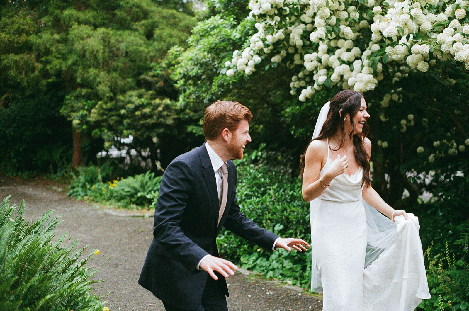 Bride and groom laughing and running through the garden, with white blossoms in the background at Parsons Gardens.