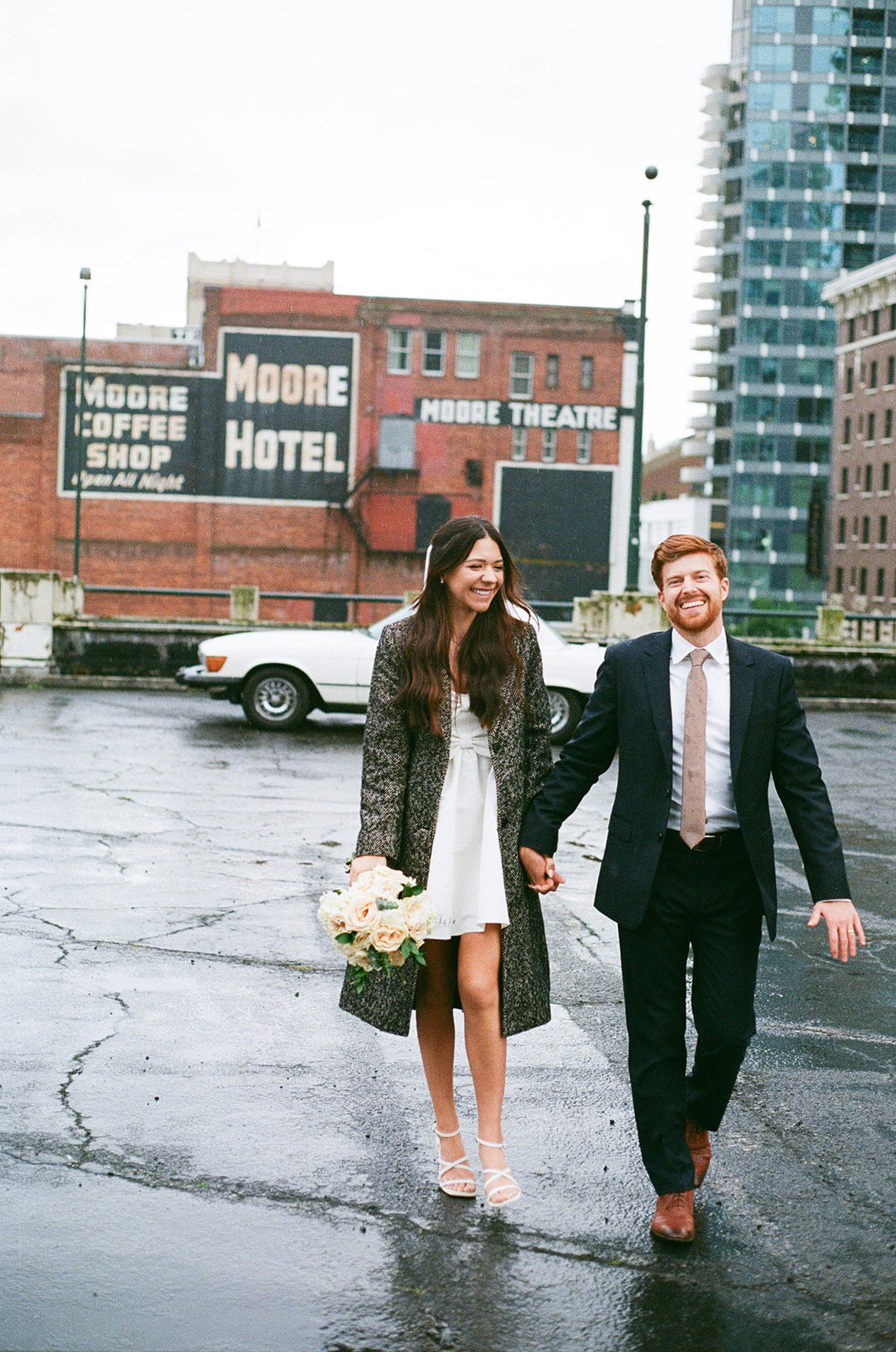 Bride and groom walking hand-in-hand in front of the Moore Theatre, with the bride wearing a long coat and holding her bouquet.