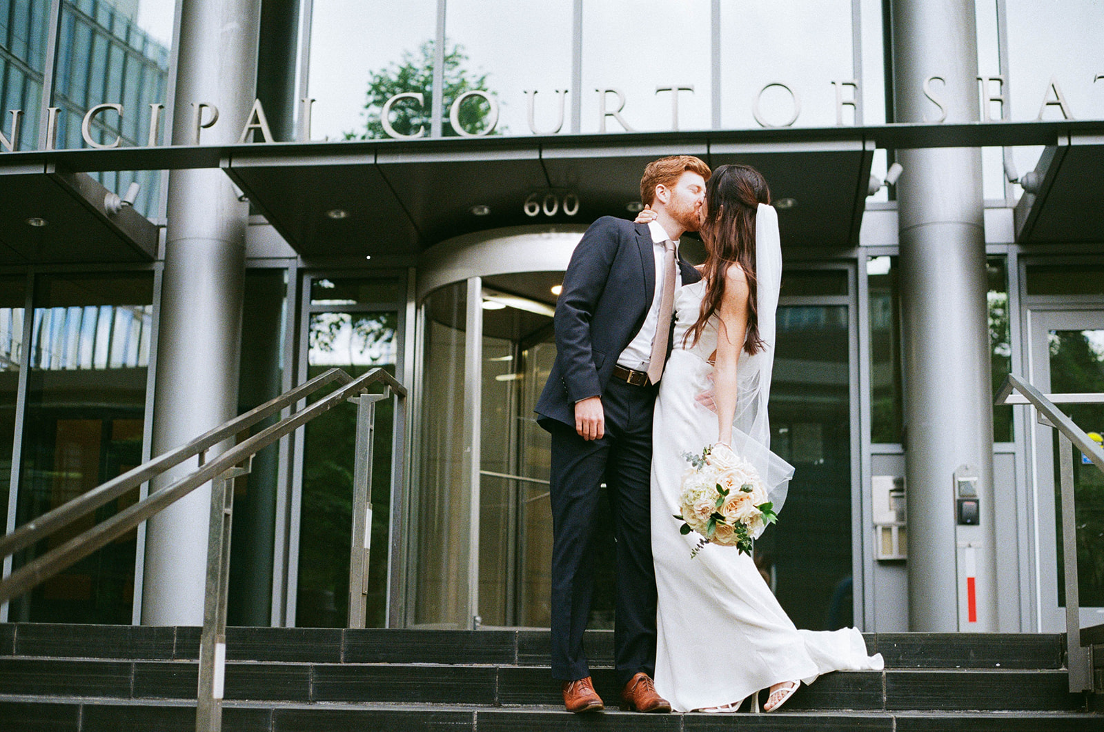 Bride and groom share a kiss on the steps of the Seattle Municipal Court, with the bride holding her bouquet.