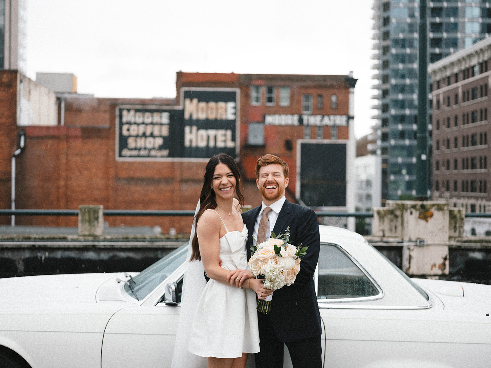 Bride and groom laughing together in front of a vintage white car with Seattle’s Moore Hotel in the background.