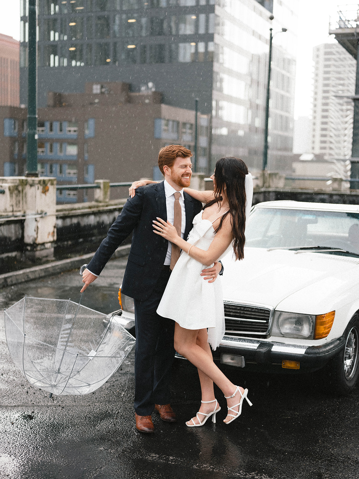 Bride and groom smiling under a light rain, standing in front of a vintage white car with a clear umbrella resting on the ground