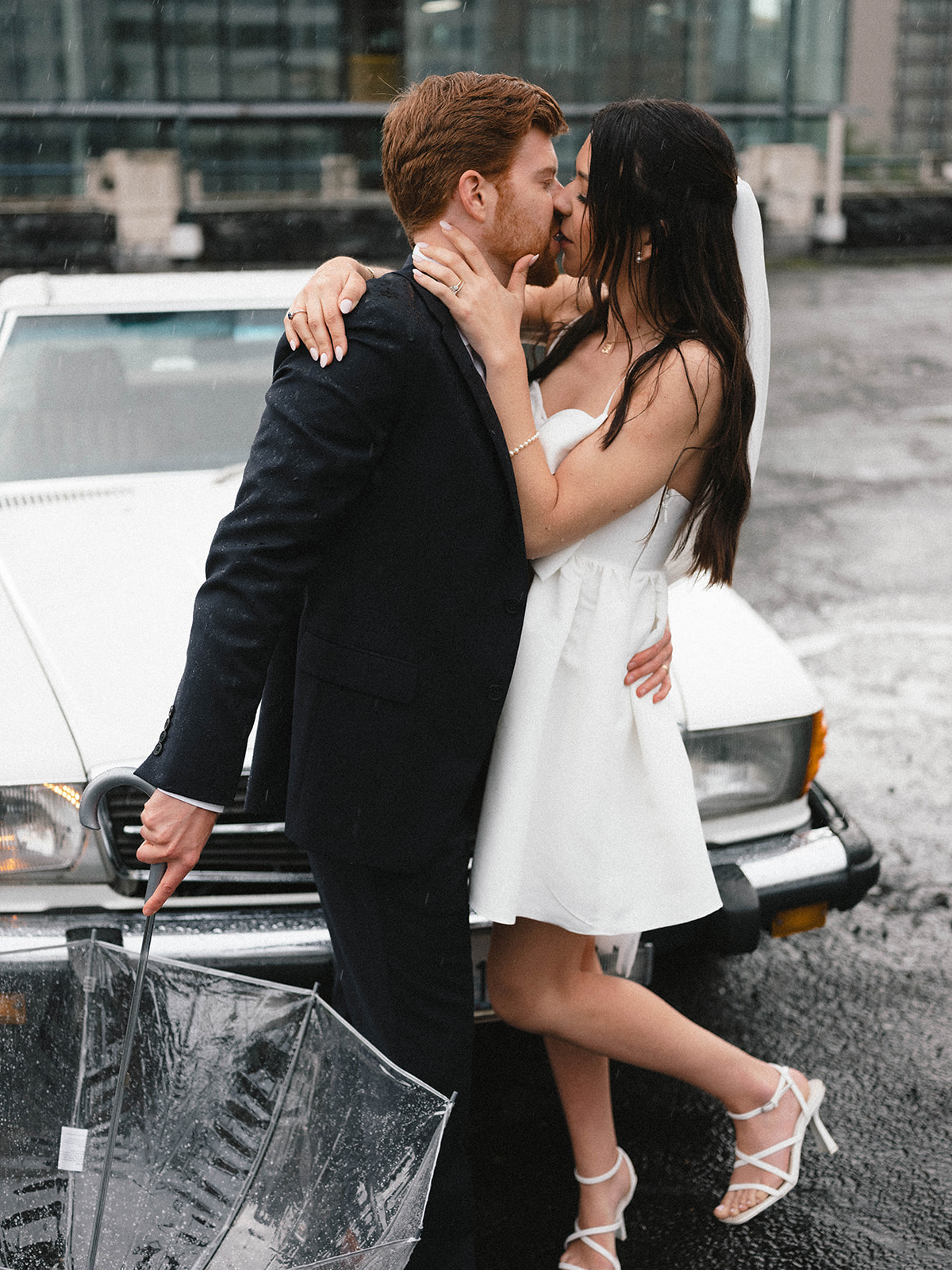 Bride and groom sharing a kiss in the rain in front of a vintage white car, with the groom holding a clear umbrella.