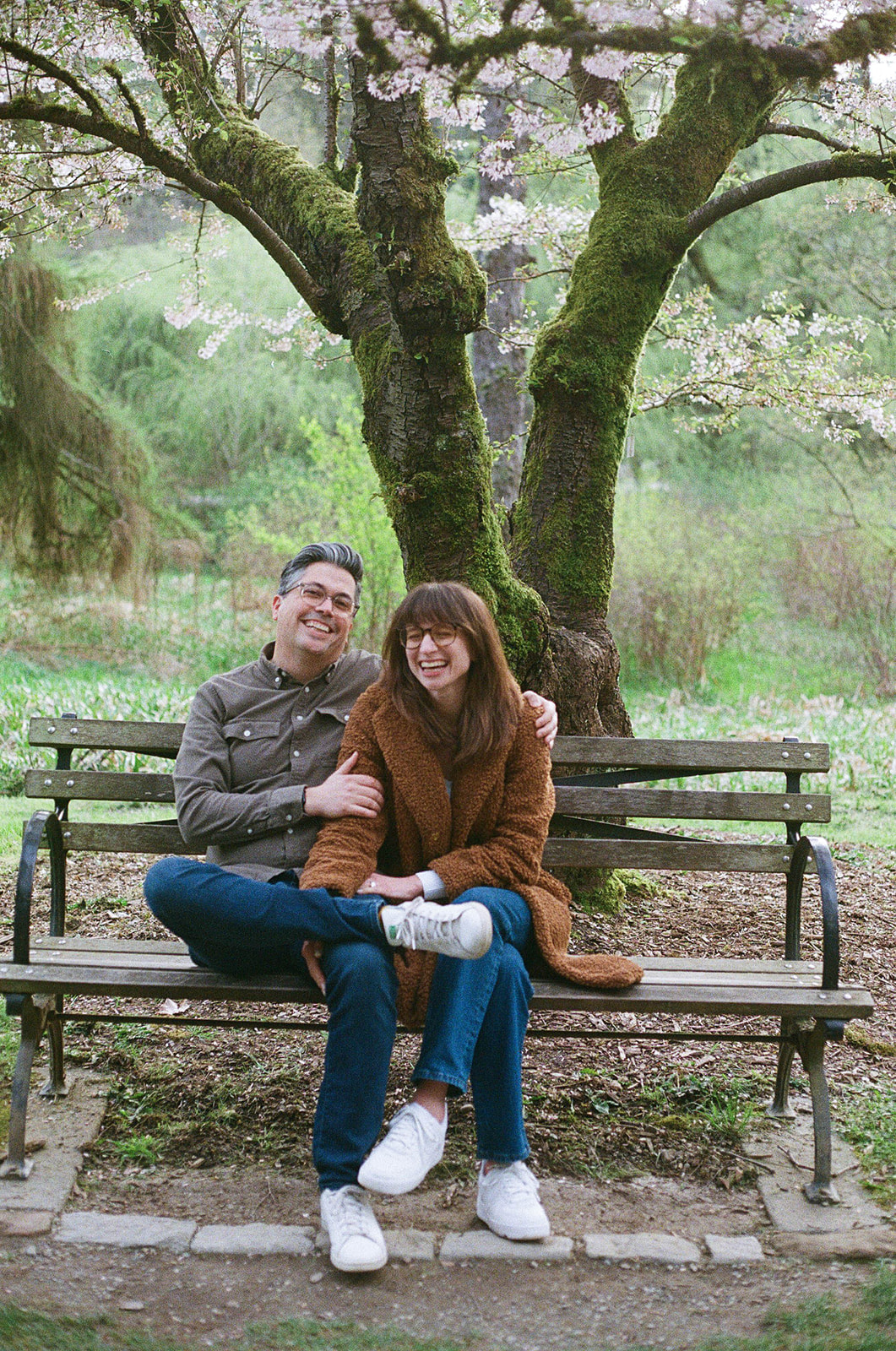 Charlotte and Grant sitting on a bench beneath a mossy tree, laughing together at Washington Park Arboretum.