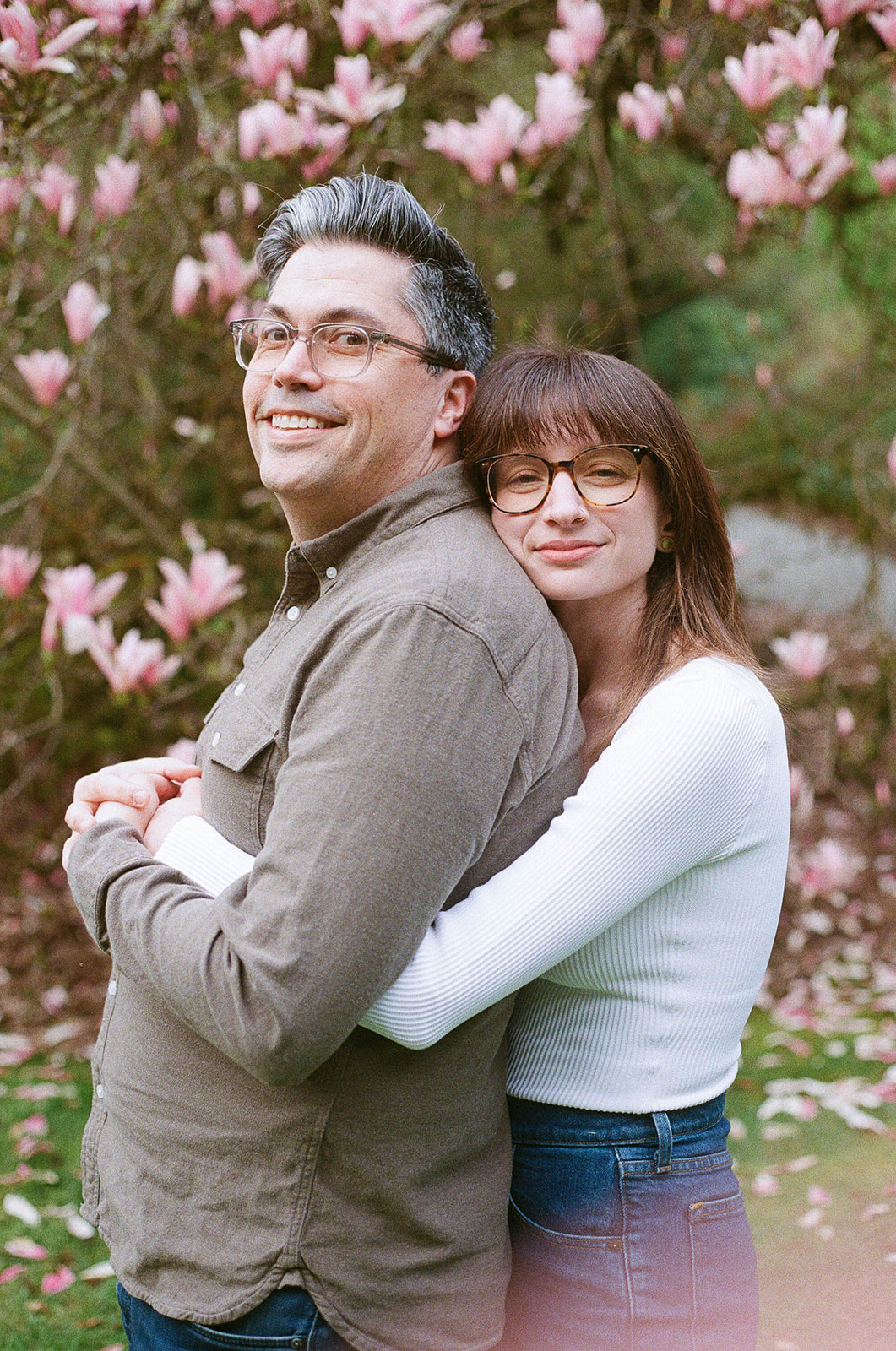 Charlotte and Grant smiling and embracing in front of a blooming magnolia tree at Washington Park Arboretum.