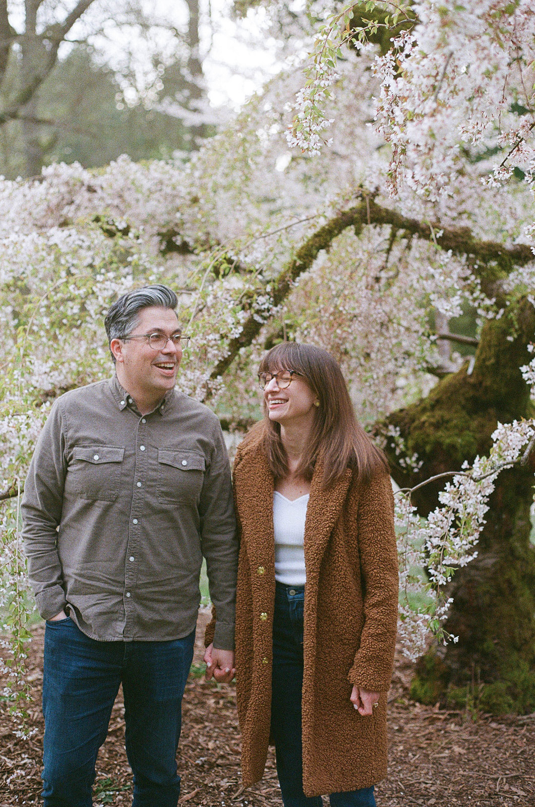 Charlotte and Grant walking hand-in-hand under a cherry blossom tree, sharing a lighthearted moment.