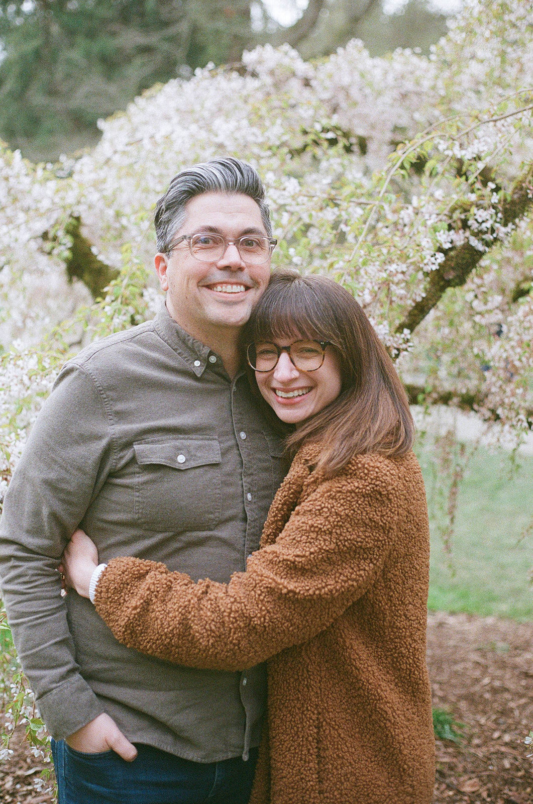 Charlotte and Grant embracing and smiling in front of a blooming cherry blossom tree.