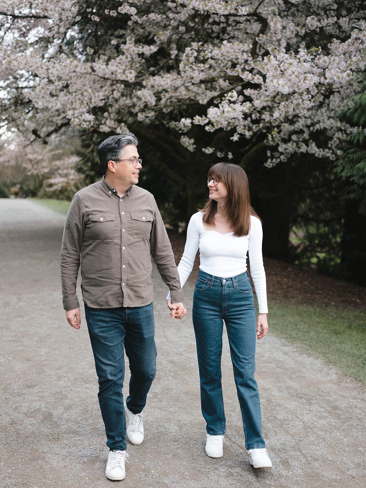 Charlotte and Grant walking hand in hand down a path lined with cherry blossoms at the Washington Park Arboretum.