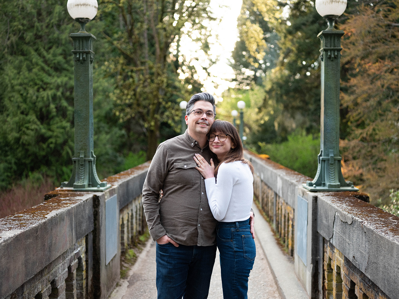 Charlotte and Grant walking hand-in-hand on a bridge surrounded by trees.