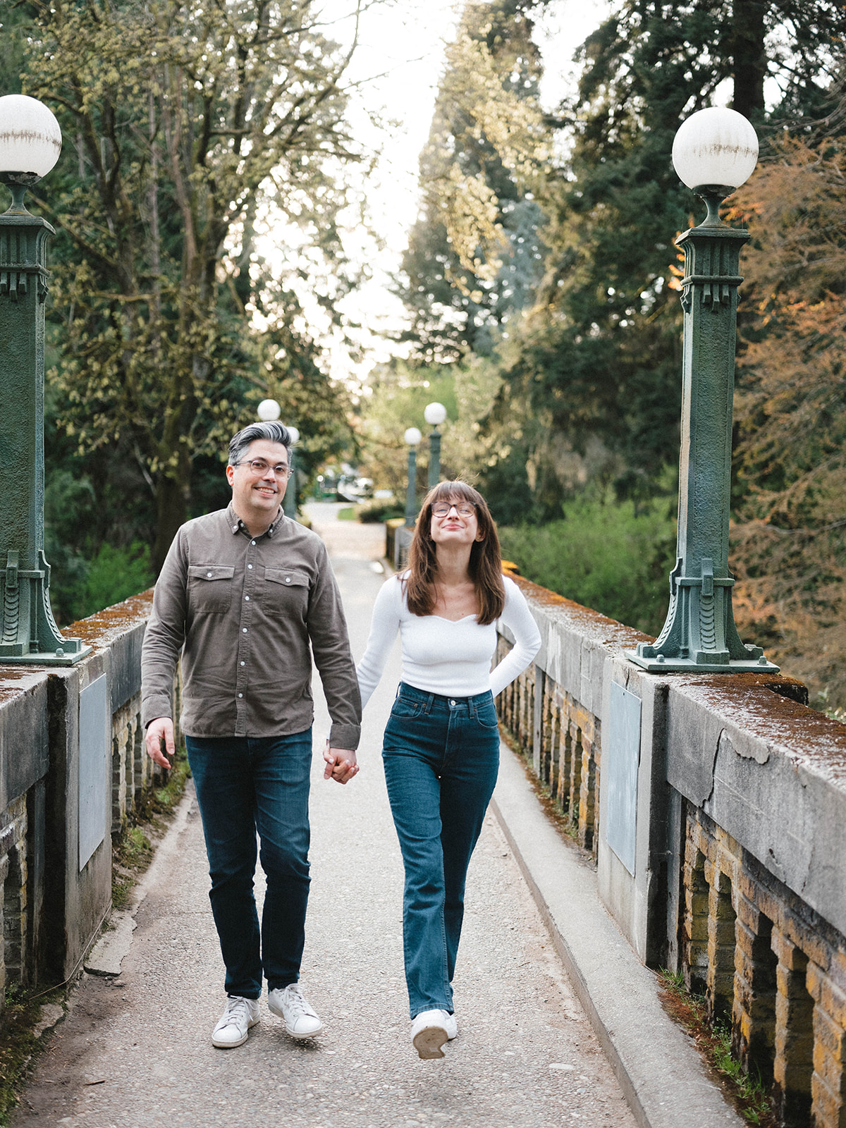 Charlotte and Grant walking hand-in-hand on a bridge surrounded by trees.