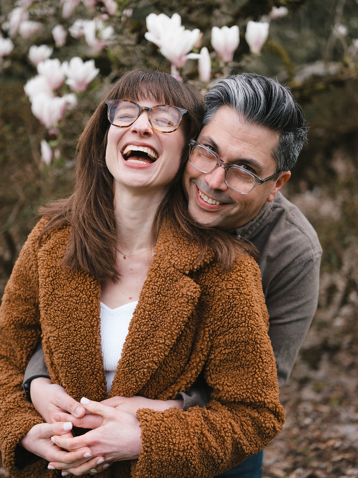 Charlotte and Grant laughing together while embracing in front of blooming magnolia trees.