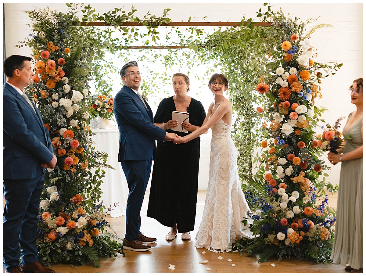 Bride and groom hold hands under a floral arch during their wedding ceremony.