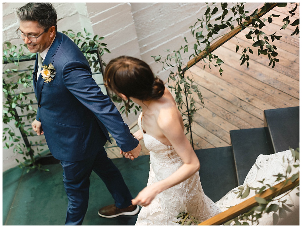 Bride and groom walking up a staircase draped with greenery, holding hands after their wedding ceremony.