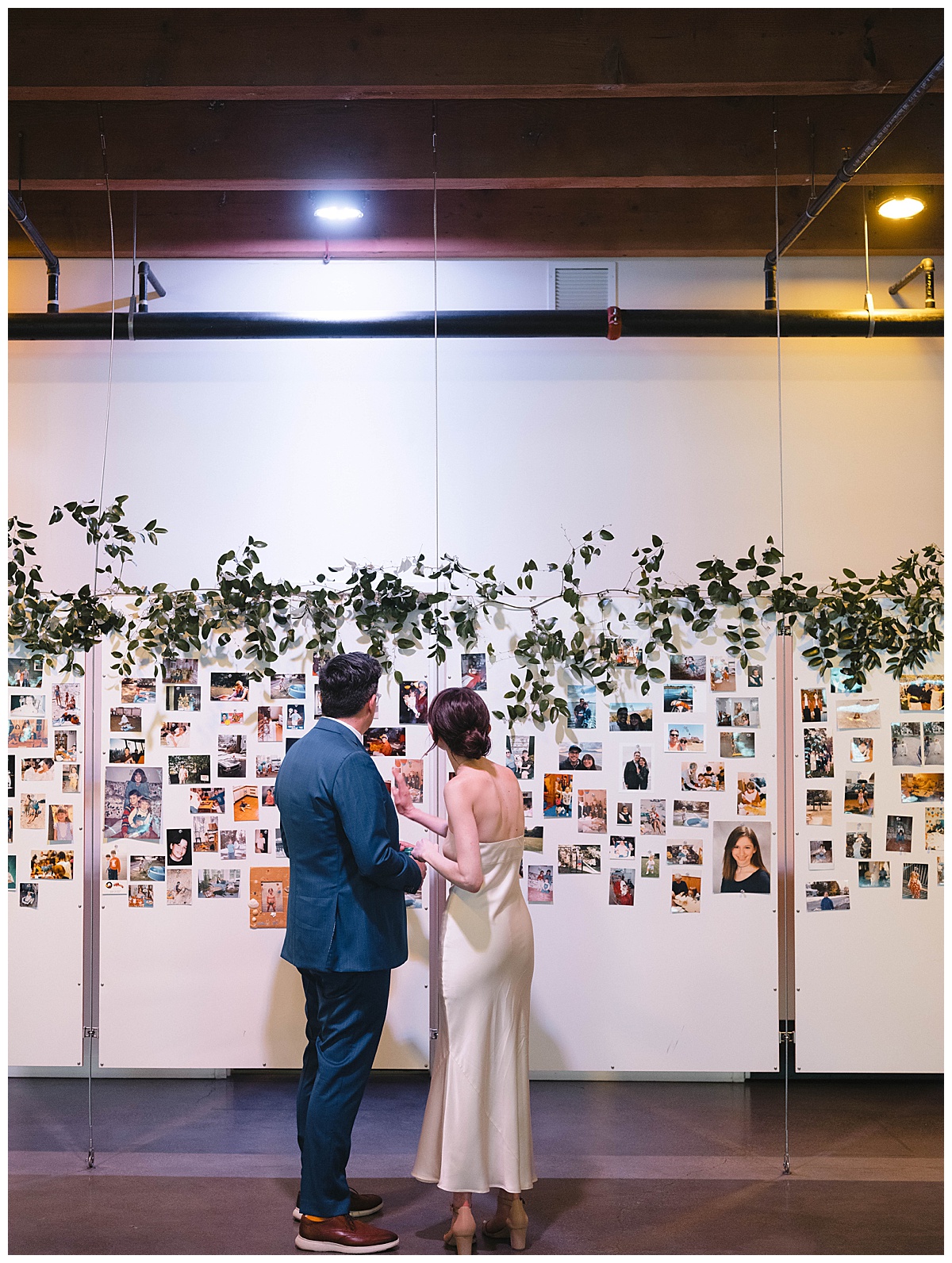 Bride and groom admire a photo wall filled with memories, decorated with greenery.