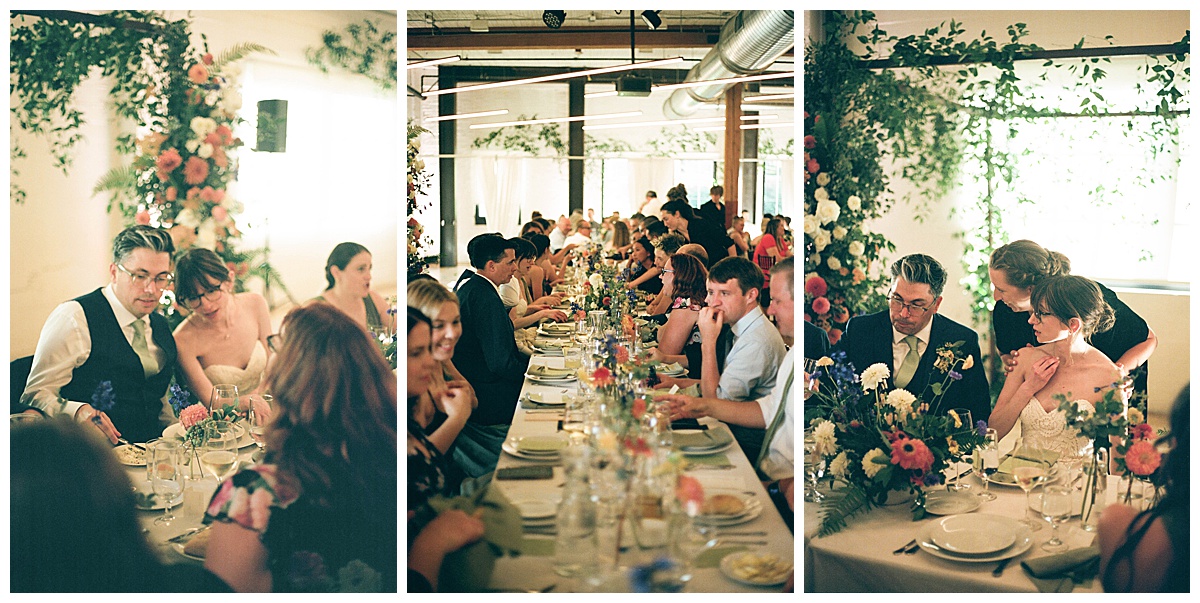 Bride and groom sitting among guests, enjoying their reception dinner at a long, flower-filled table.