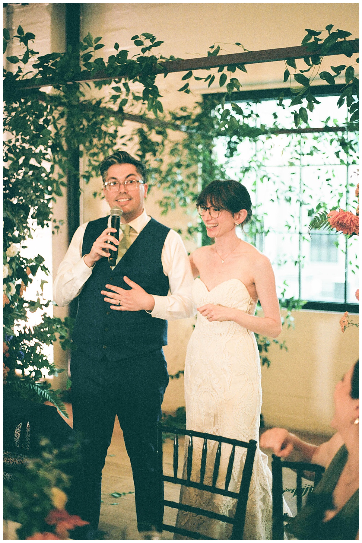 Groom giving a speech during the wedding reception, standing beside the bride with a microphone.