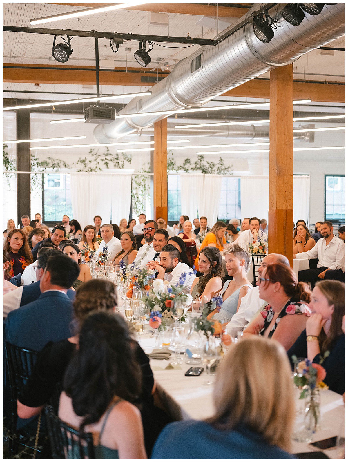 The bride and groom share a quiet moment with guests during the reception dinner, with the bride seated at a long dining table filled with flowers and guests in the background.