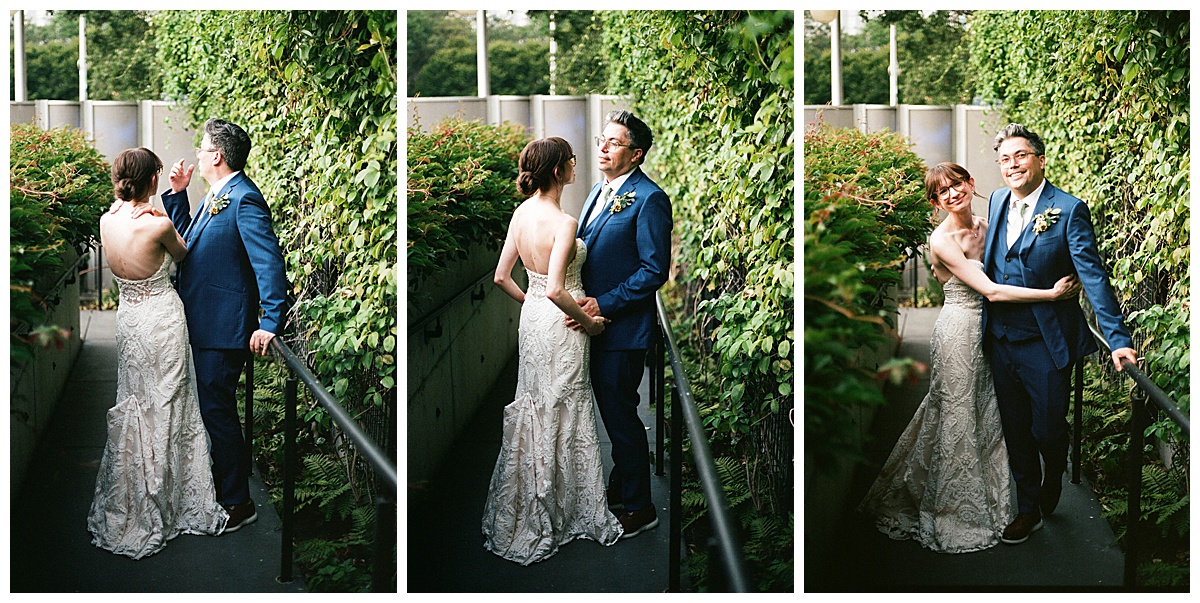 The bride and groom pose together outdoors, surrounded by greenery on a walkway, sharing affectionate glances and smiles.