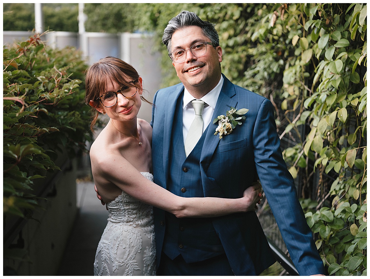 The bride and groom stand close together, embracing under a canopy of greenery, smiling and celebrating their special day.