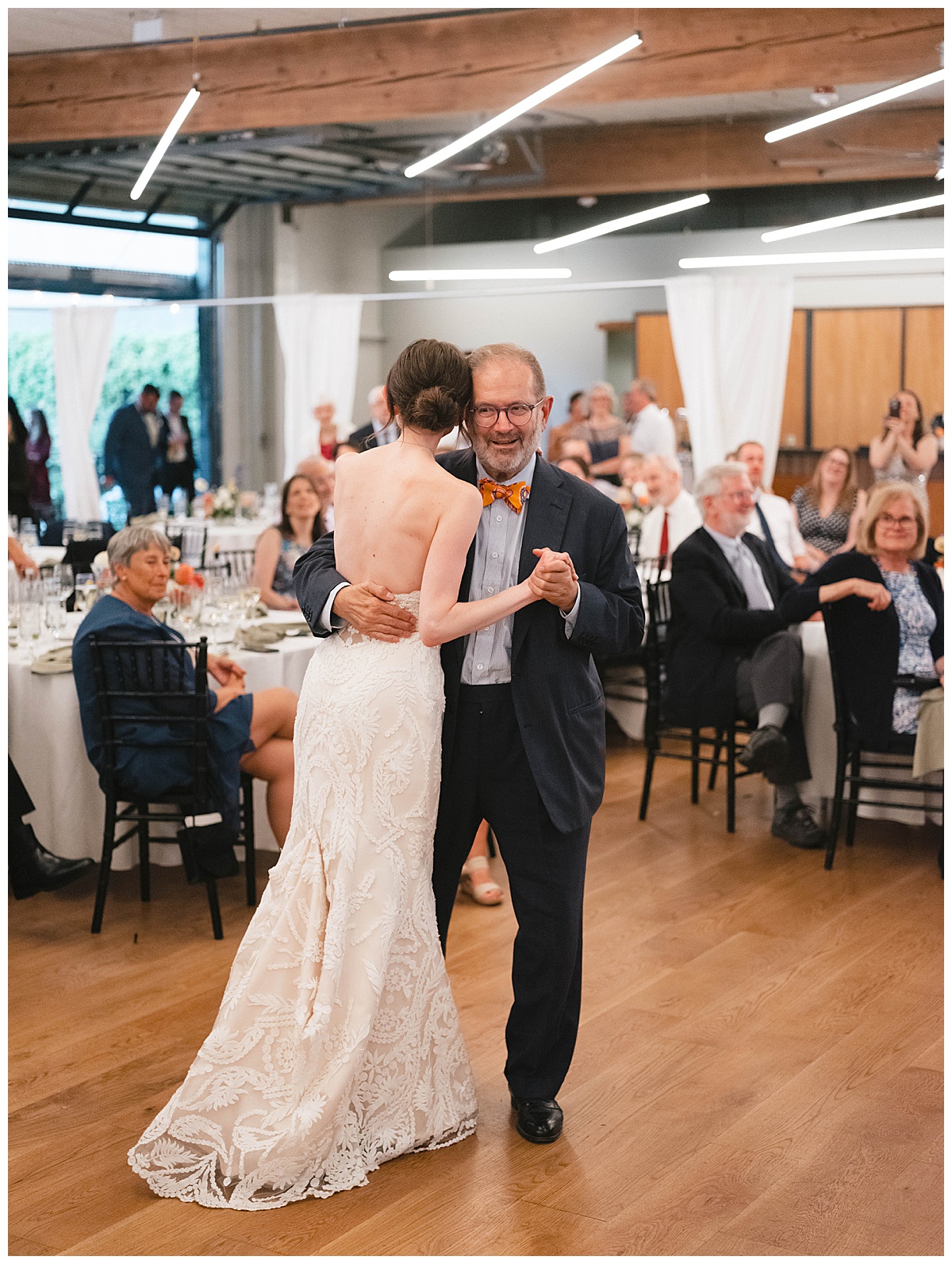 Bride dances with her father, smiling warmly as guests watch.