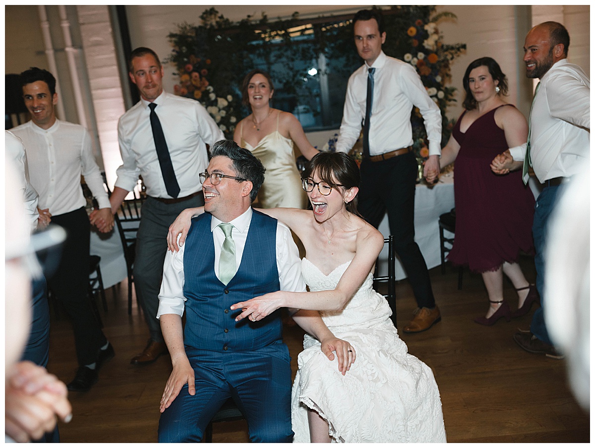 The bride and groom laugh together while seated at the center of a group dancing around them.