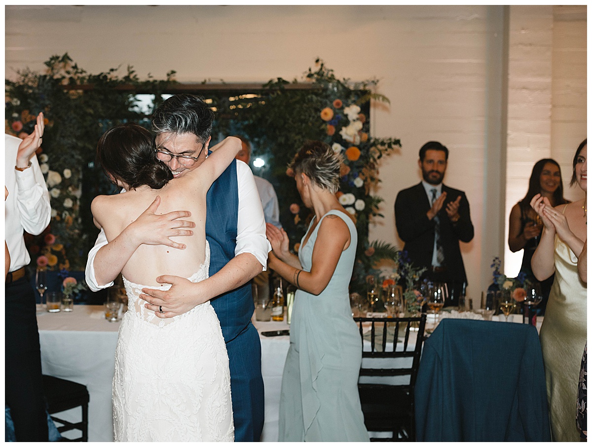 The couple shares an intimate moment during the speeches, embraced in a heartfelt hug while guests cheer around them.