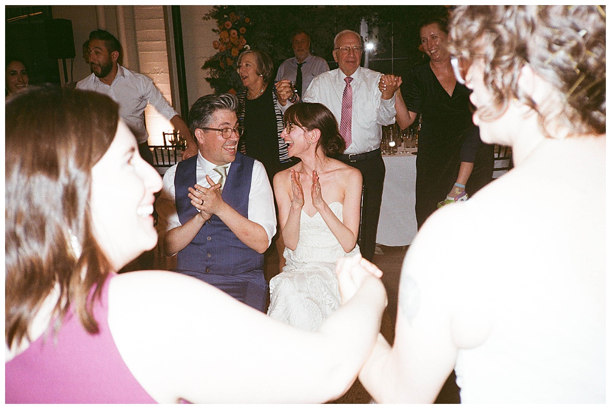 The couple sits at the center of a celebratory dance, surrounded by friends holding hands in a traditional circle dance.