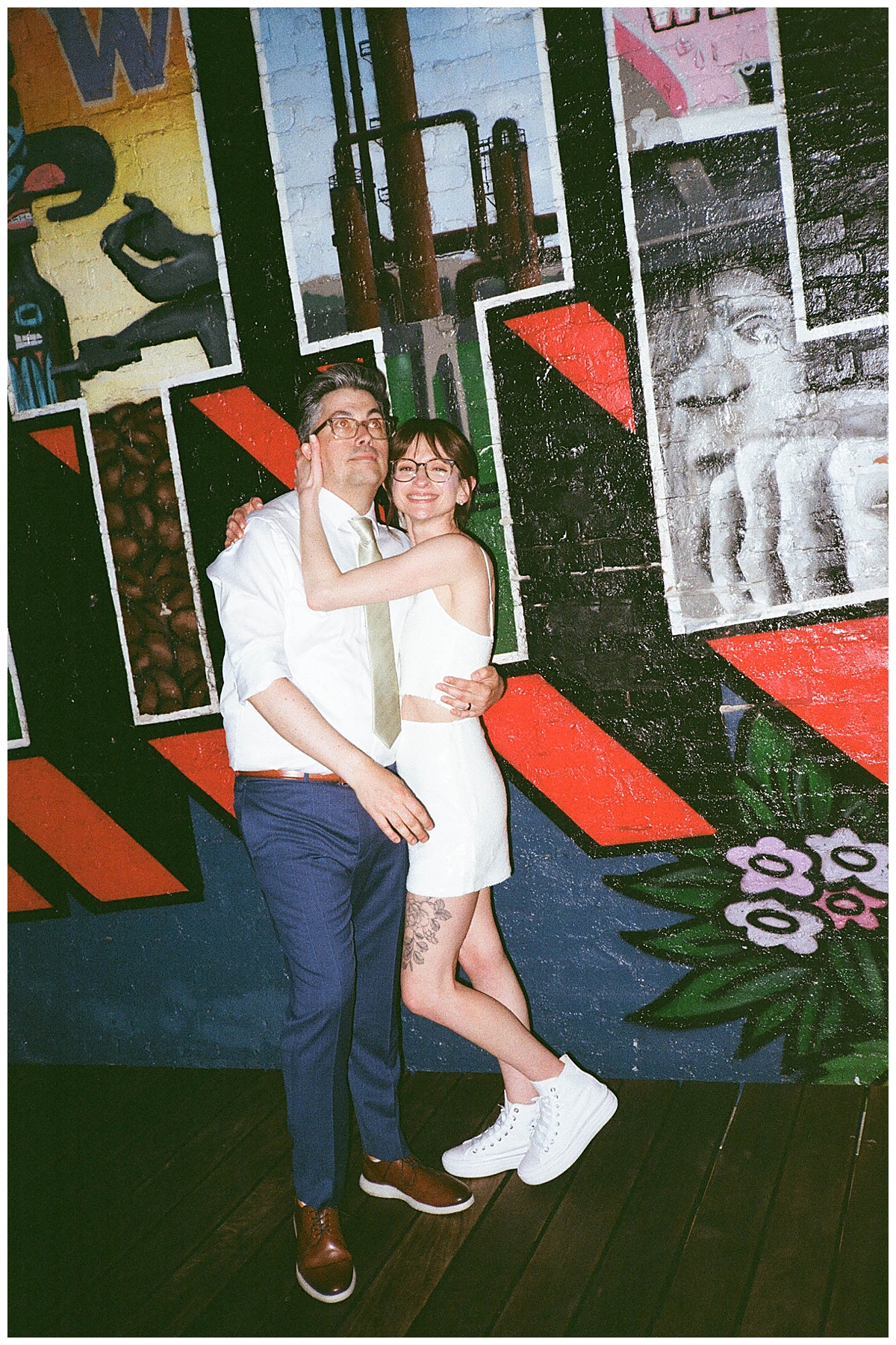 The couple smiles and embraces against a colorful mural backdrop.