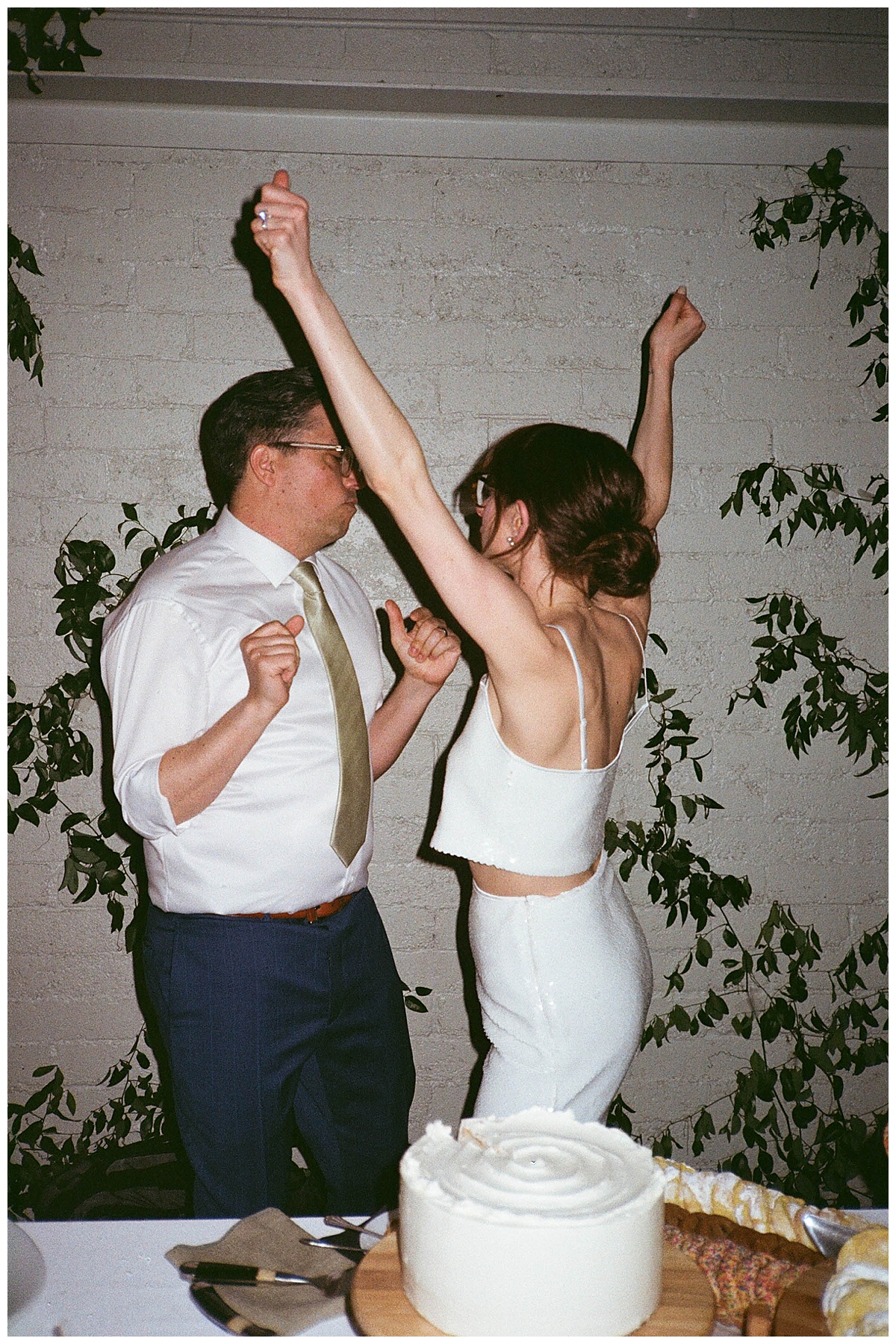 Bride raises her arms in celebration while the couple dances beside their wedding cake.